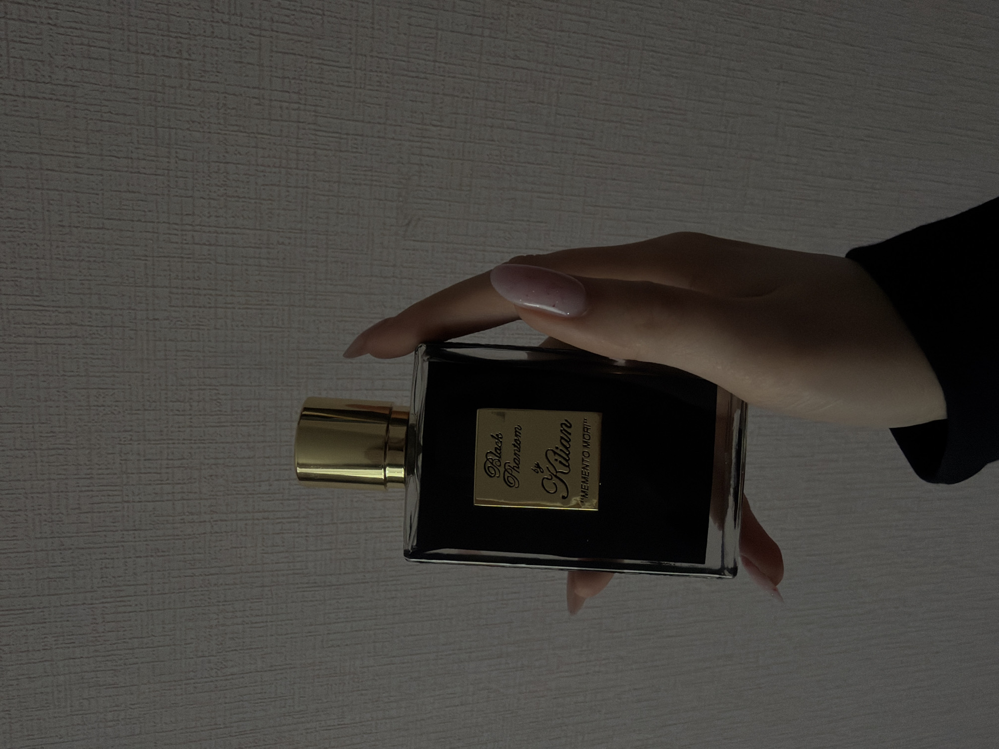 A hand is holding a rectangular black glass bottle with a gold cap and a gold label with the words "Black Phantom by Kilian 'MEMENTO MORI'" on it. The background is a textured gray wall. The location is not discernible from the image.