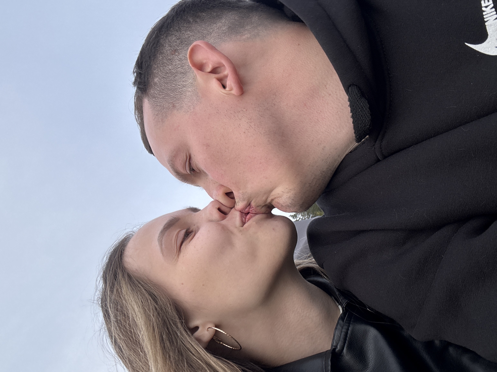 A close-up, vertically oriented photograph captures a man and a woman kissing outdoors. The man is wearing a black hooded sweatshirt with a white Nike logo partially visible. The woman is wearing a black leather jacket and a gold hoop earring. The background is overcast with a light-colored sky. The photograph was taken outdoors in the city of Ольховка, Russia.