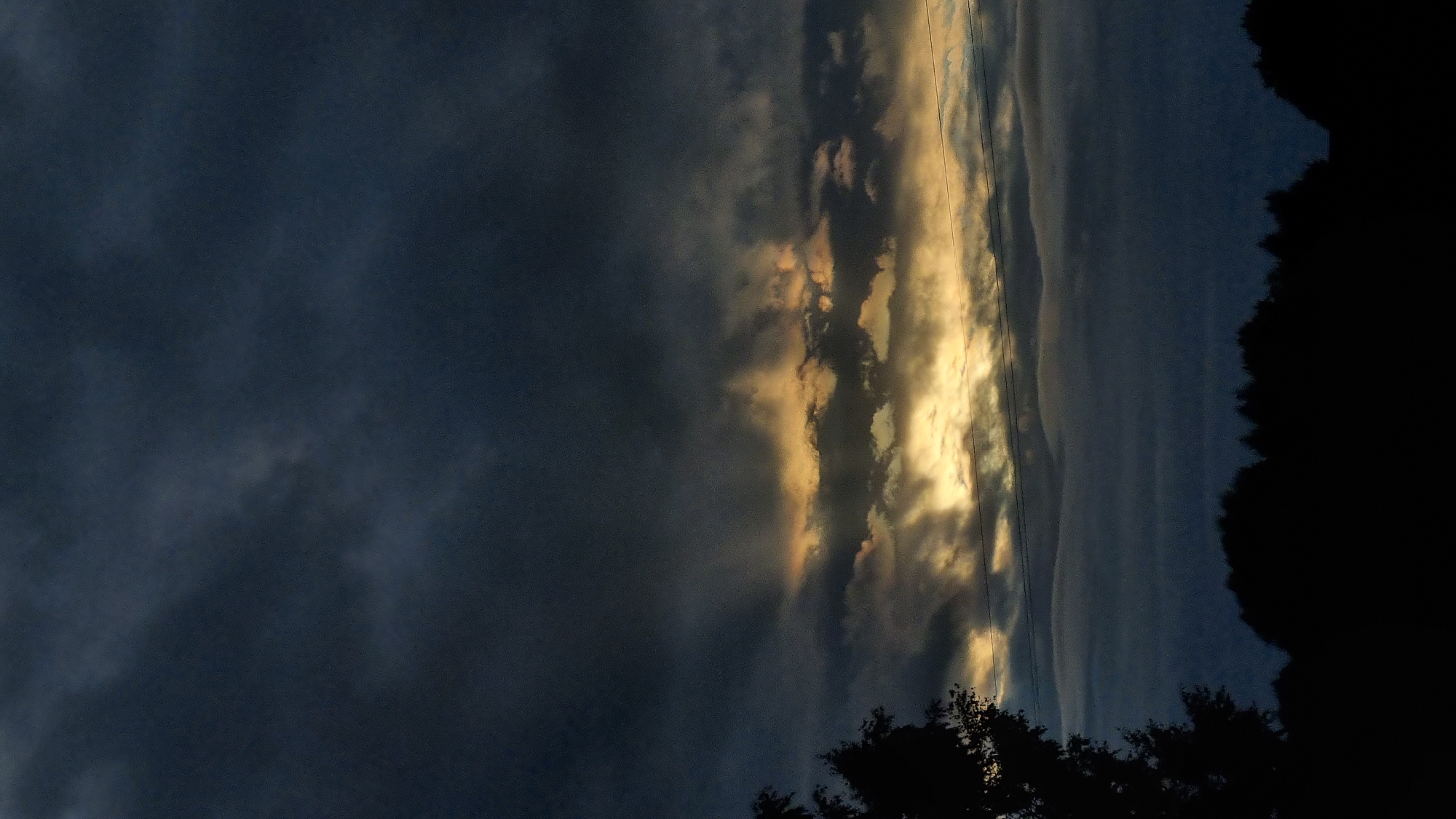 The image depicts a stormy sky over a silhouetted treeline, possibly in the city of Vysokoe, Russia. The sky is dominated by dark, heavy clouds, with a break in the clouds revealing a golden, illuminated layer, suggesting either a sunset or sunrise behind the storm. A single tree is visible on the left side of the frame, also in silhouette. Power lines stretch horizontally across the middle ground. The overall impression is one of dramatic weather, with the natural landscape rendered in stark contrast against the illuminated sky.