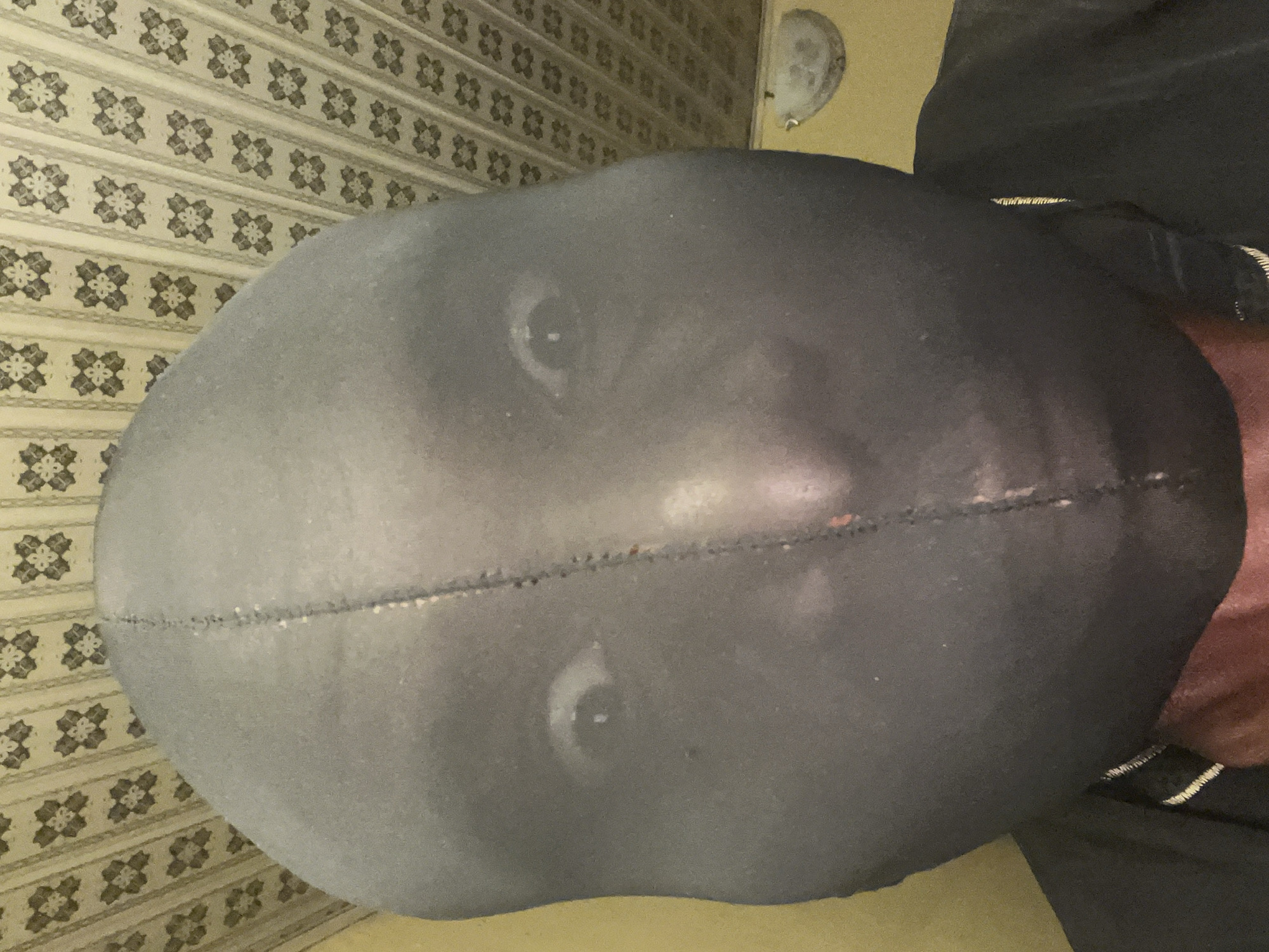 A person wearing a black balaclava is seen from the top, looking towards the camera. The background includes a patterned wallpaper and a glimpse of clothing near the person's shoulder. The image was taken indoors. There is no other information suggesting this is in Gura Top, Nigeria.