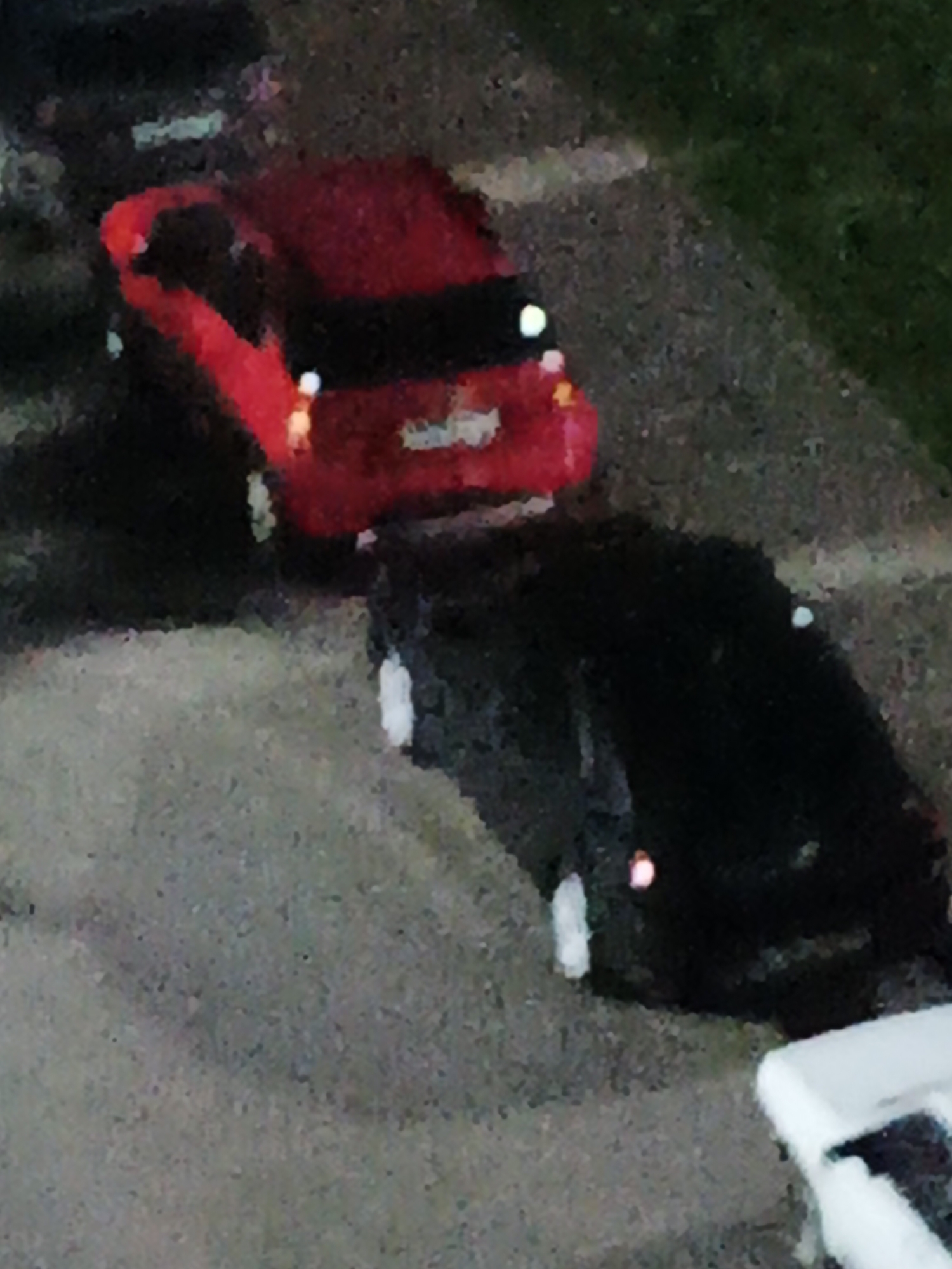 This is a low-resolution, aerial, nighttime photograph of a residential area in Kazan, Russia. The image depicts two parked cars, one red and one black, on a paved surface that appears to be a driveway or parking lot. The red car is in the foreground, and the black car is behind it. A small portion of a white object, possibly another vehicle or structure, is visible in the lower right corner. The surrounding environment includes asphalt, grass, and what appears to be a sidewalk or curb. The lighting conditions suggest it is dark, with artificial light sources casting reflections on the vehicles. There are no people visible in the image.