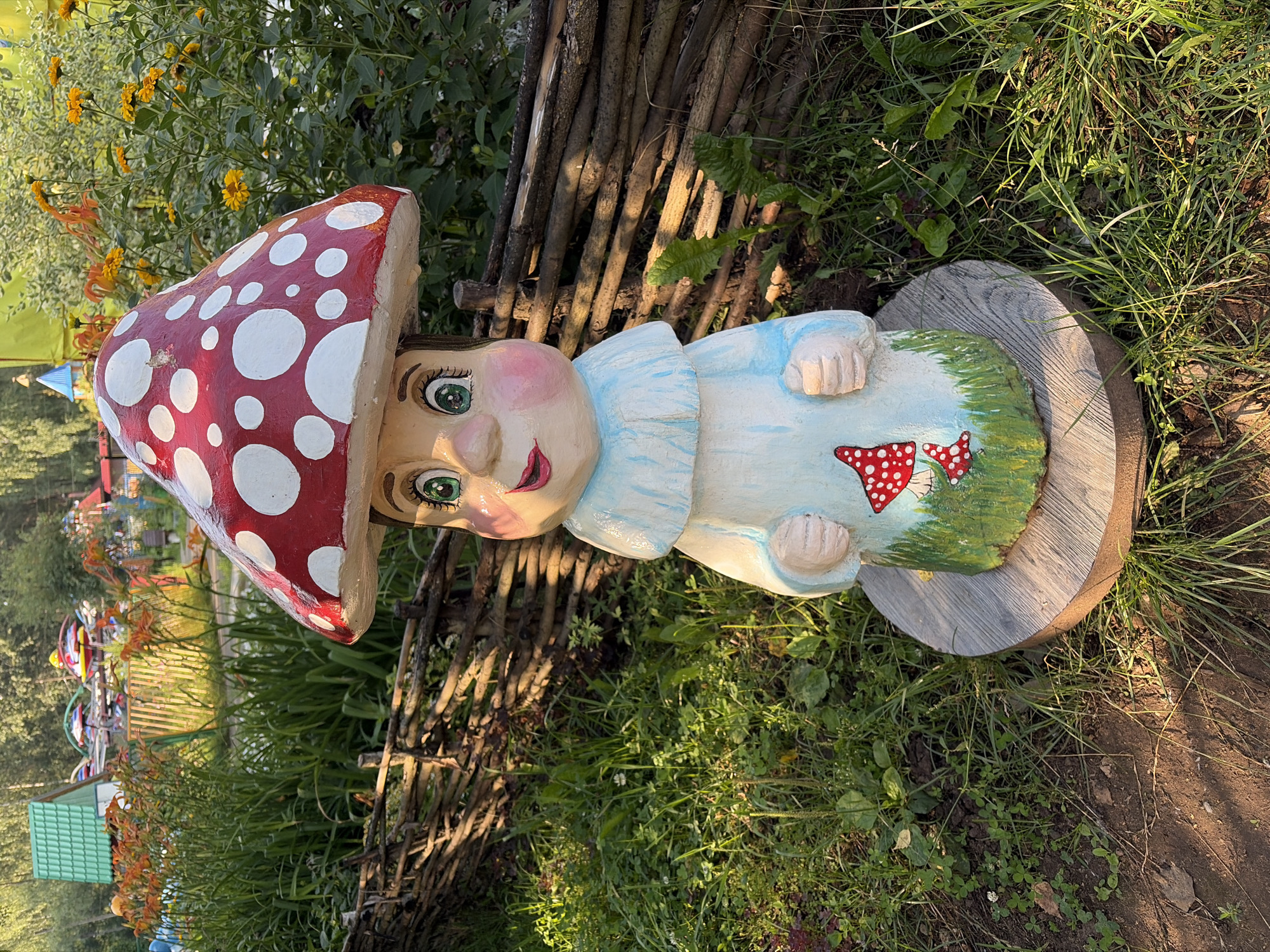 A gnome-like mushroom statue stands in a park in Chaikovsky, Russia. The statue, painted to resemble a red and white spotted mushroom, has a human-like face with green eyes, rosy cheeks, and red lips. It wears a light blue tunic with a ruffled collar and has a painting of two smaller mushrooms on the front of its lower body, which is painted to look like grass. The gnome is mounted on a light gray, circular base. Behind the statue, a rustic wooden fence made of woven branches separates the immediate area from a grassy lawn and a flower bed with orange and yellow blossoms. In the background, several amusement park rides, including a Ferris wheel and a roller coaster, are visible, suggesting the park is a recreational area. The scene is illuminated by sunlight, casting shadows on the ground.