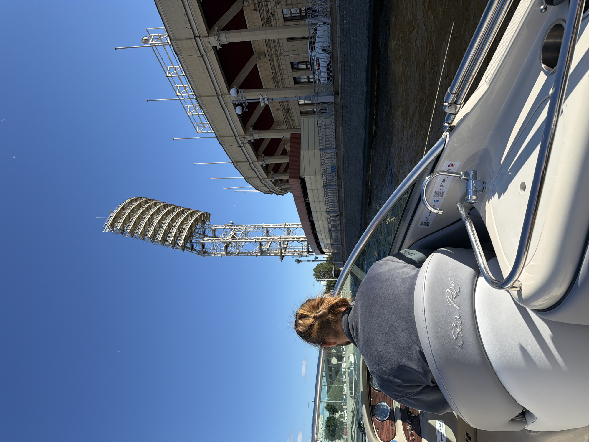 A person with blonde hair tied in a bun is driving a boat on a river, viewed from behind. The boat is a Sea Ray. In the background, a large stadium with bright floodlights and a curved facade is visible. The sky is clear and blue. The water is dark and rippling. Buildings and trees are also seen along the riverbank.