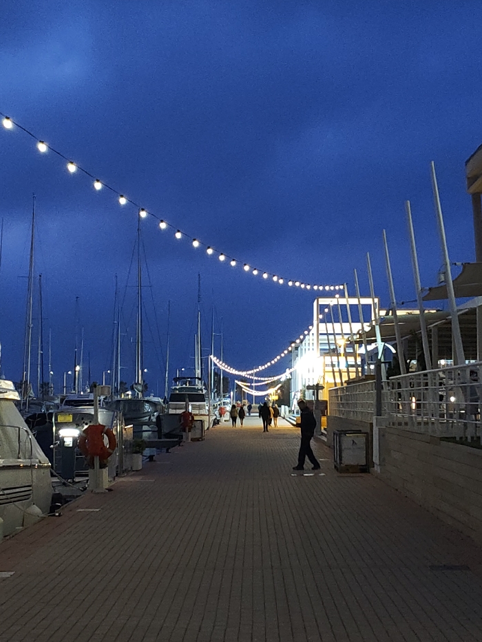 The image captures an evening scene at the marina in Dénia, Spain. A brick-paved promenade, composed of rectangular pavers arranged in a linear pattern, extends from the foreground into the background. To the left of the promenade, numerous yachts and motorboats are docked, their tall masts visible against the dark blue twilight sky. One orange lifebuoy is affixed to a white post on the quay in the mid-ground. To the right, a series of modern buildings with illuminated interiors and outdoor terrace areas featuring white vertical support poles and awnings line the promenade. Overhead, two prominent strings of warm-toned spherical lights are suspended, forming graceful curves that provide ambient illumination along the pathway. Approximately ten individuals are distributed along the promenade, walking in various directions. A person in dark clothing is visible in the mid-ground, walking towards the right side of the frame. A light-colored, rectangular container or bin is positioned on the right edge of the promenade. The sky above is a uniform deep blue, indicative of a clear evening or night.