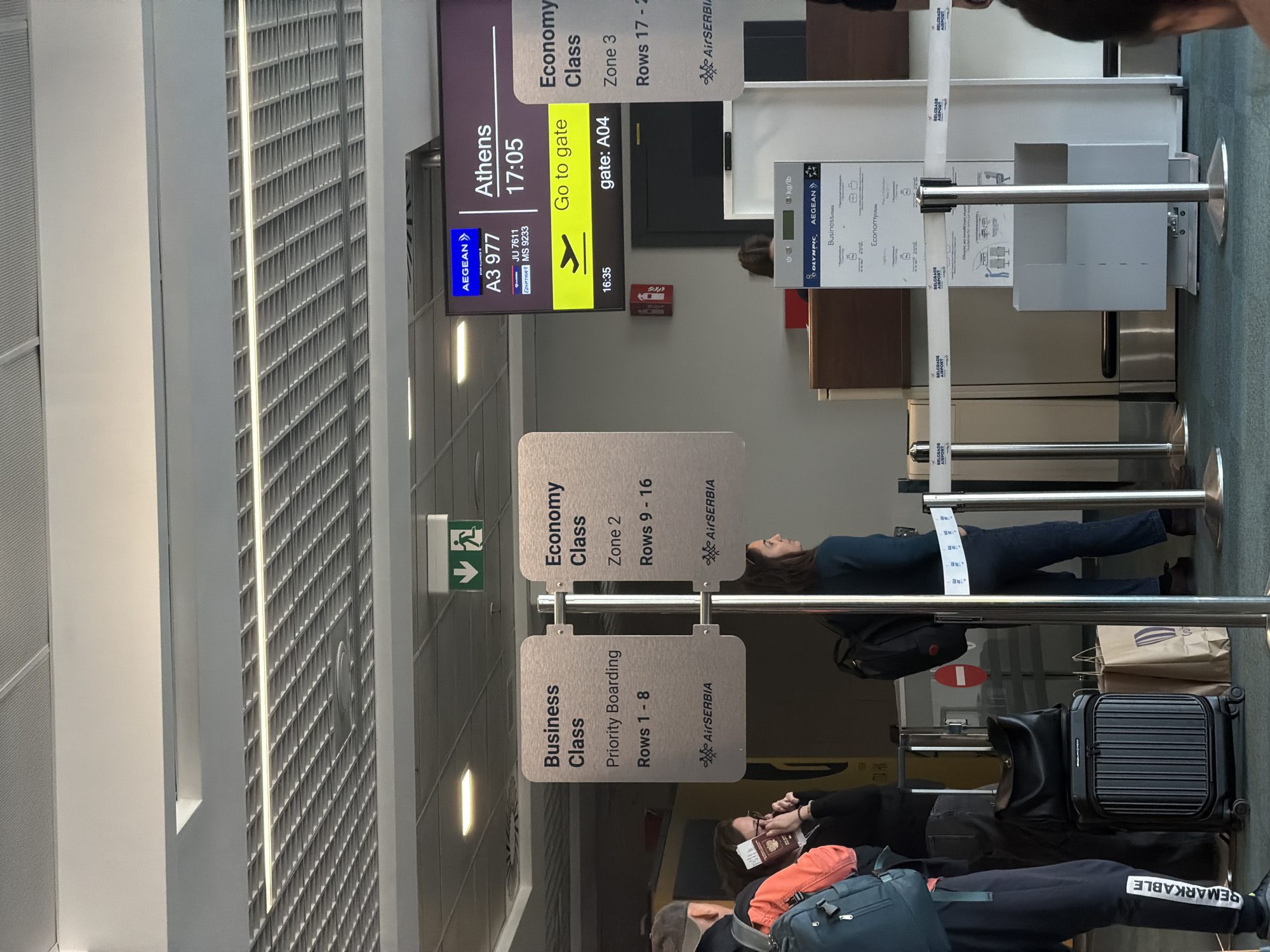 This media file captures a scene at a boarding gate in Belgrade Airport, bustling with passengers waiting to board a flight. The setting is an indoor airport terminal, featuring a patterned ceiling, artificial lighting, and a carpeted floor.
A large digital screen prominently displays flight information for a flight to "Athens" on "AEGEAN" flight "A3 977", with code shares "JU 7611" (Air Serbia) and "MS 9233" (Egyptair). The scheduled departure time is "17:05", and the screen advises "Go to gate" for "gate: A04", with a time of "16:35" displayed below, indicating it's late afternoon and boarding is likely underway or about to begin.
Hanging signs categorize boarding zones: "Business Class" (Priority Boarding, Rows 1-8), "Economy Class Zone 2" (Rows 9-16), and "Economy Class Zone 3" (Rows 17-2?), all bearing the "AirSERBIA" logo. Further back, another information stand from "BELGRADE AIRPORT" displays baggage guidelines in "kg/lb" for "Business-class" and "Economy-class", featuring "OLYMPIC," "AEGEAN," and "AirSERBIA" logos.
Several people are visible in various states of waiting. In the foreground, an individual wearing dark sweatpants with "REMARKABLE" printed on the leg holds a passport or phone, positioned next to a blue backpack and a black rolling suitcase. Another person in a dark blue top and jeans stands further back, looking up towards the signs. The overall atmosphere appears calm, with passengers seemingly engaged with their devices or waiting patiently. An exit sign and a fire alarm are visible on the walls.