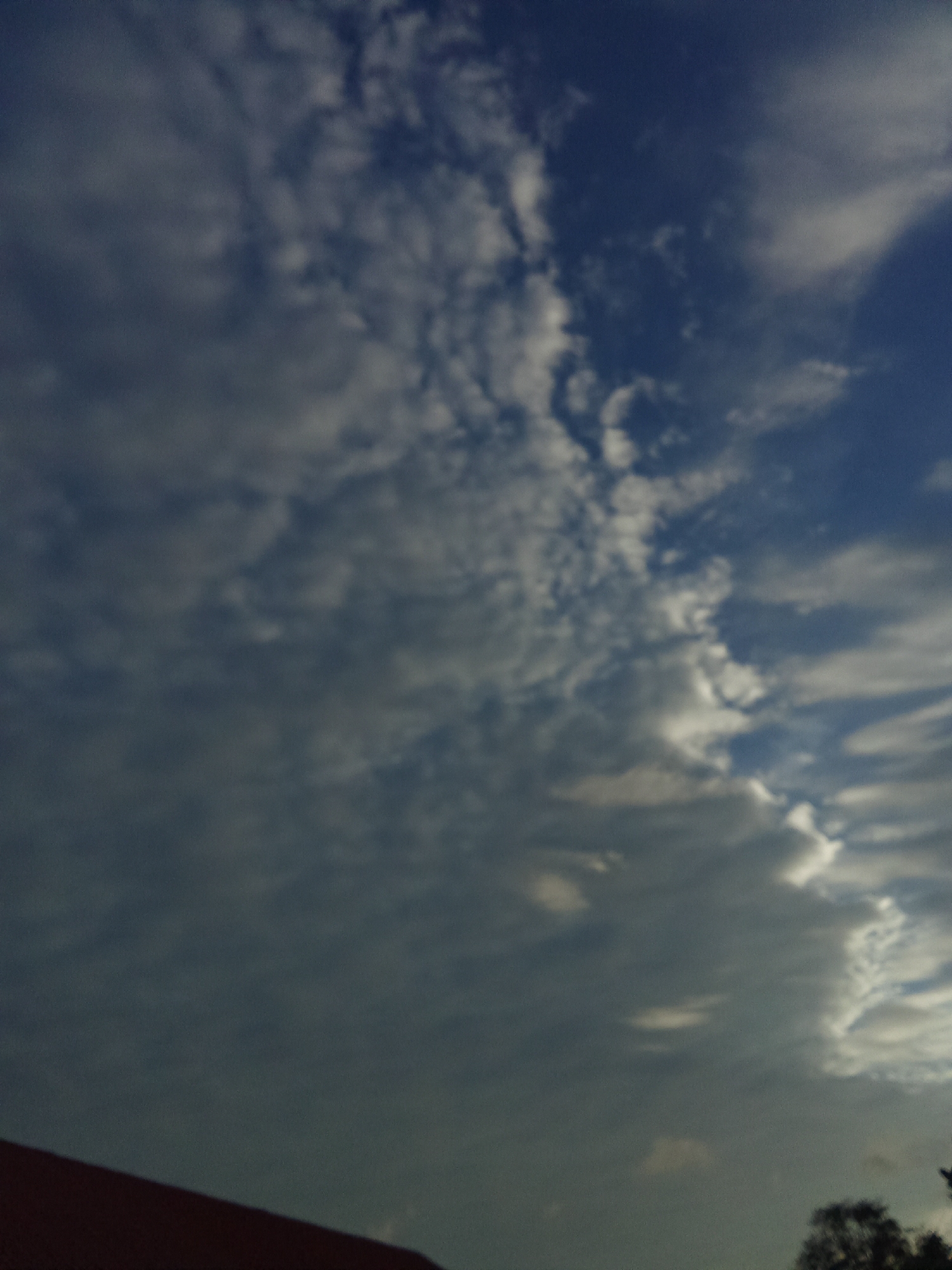 The image shows a cloudy sky in Juan Griego, Venezuela. The sky transitions from a darker, overcast gray on the left side to a lighter blue with white clouds on the right. A small portion of a red roof is visible in the lower left corner, and some trees are discernible in the lower right corner. The image is slightly out of focus.