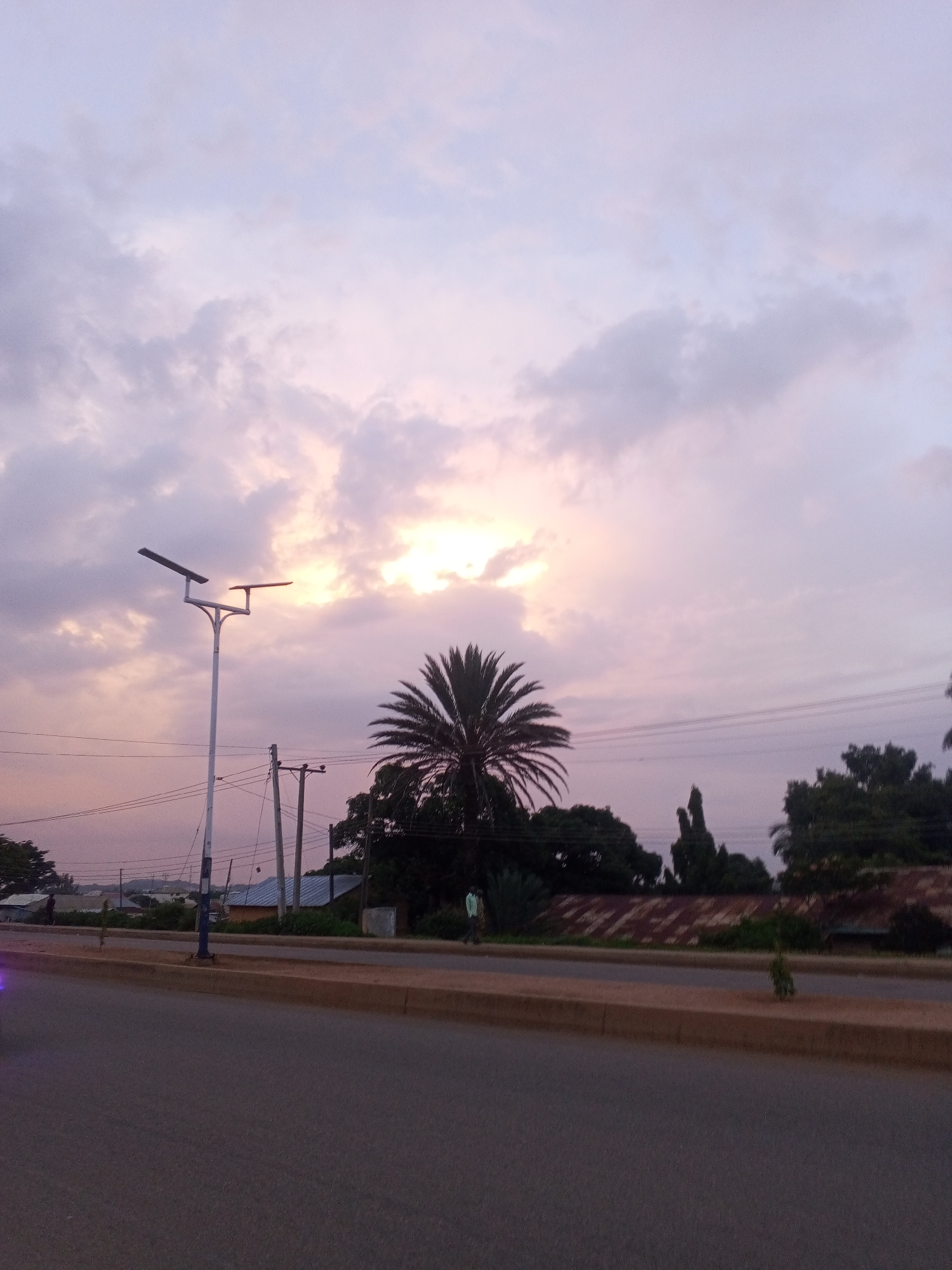 The image depicts a street scene in Bukuru, Nigeria, during what appears to be twilight or early morning. The sky is filled with soft, muted clouds, tinged with pink and purple hues, with the sun faintly visible through the haze. A tall, white street light with two arms stands prominently in the foreground to the left, its solar panels angled towards the sky. Across the road, a single palm tree with a distinctive crown of fronds stands near a cluster of other trees and vegetation. In the background, there are signs of human habitation, including buildings with corrugated iron roofs and utility poles with electrical wires. A road, likely a main thoroughfare, occupies the bottom portion of the image, with a paved median and sidewalk. Two figures are visible: one in the distance on the left side of the road, near a vehicle, and another standing on the right side of the road, further back. The overall atmosphere is quiet and serene, characteristic of a transitional time of day in a residential or semi-urban area.