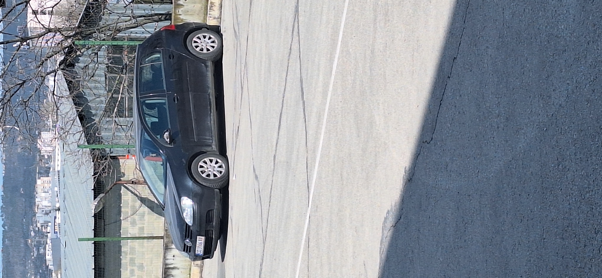 The image shows a black Volkswagen Golf parked in a paved area. The car has the license plate CD 1166 AX. The background includes a view of Varna, Bulgaria, with buildings and trees visible in the distance. A metal fence is present on the left side of the car. The paved area has lines painted on it, and there is a shadow on the right side.