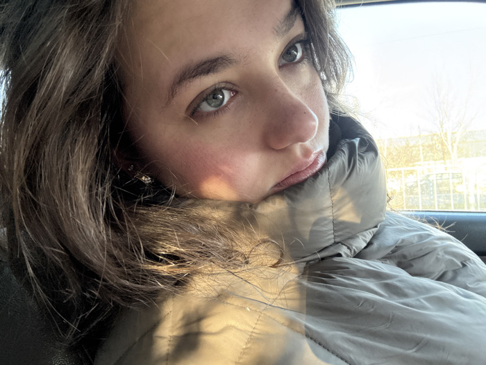 A young woman with light olive skin and wavy brown hair is shown from the chest up in a close-up. She is wearing a puffy gray jacket and seated in what appears to be a car. She gazes upwards and towards the right with green eyes. Light strikes the side of her face and jacket, casting shadows and highlights. A glimpse of a building and bare trees can be seen in the background through the car window.