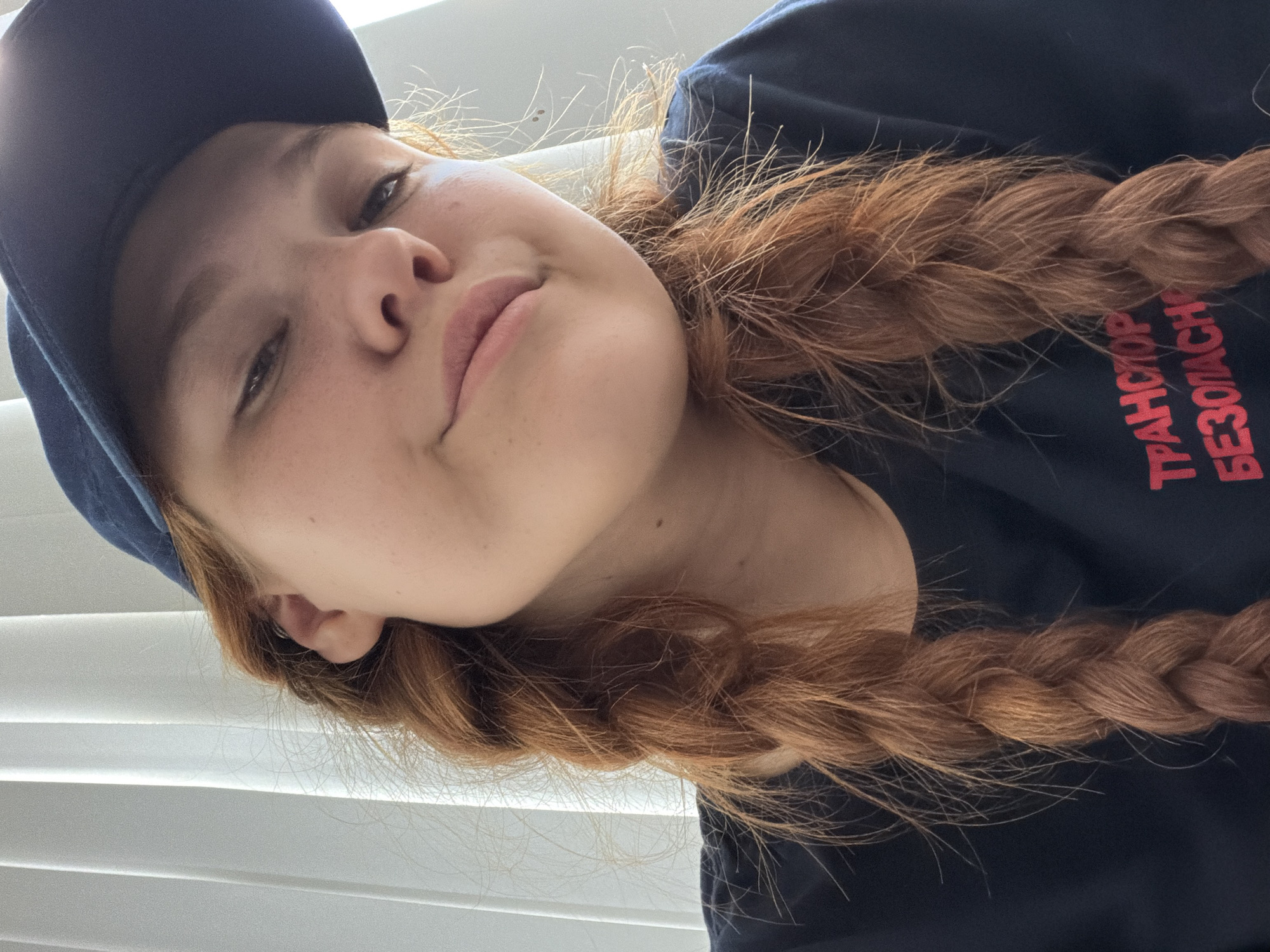 This is a portrait of a young person wearing a dark blue baseball cap and a dark blue t-shirt with red text. The person has fair skin, light freckles, and reddish-brown hair styled in a thick braid that falls over their shoulder and down their chest. Their eyes are partially closed, and they have a slight smile, conveying a relaxed or playful emotion. The background appears to be indoors, with white horizontal blinds visible, suggesting it is daytime with sunlight filtering through. The red text on the t-shirt is partially visible, with the words "ТРАНСПОРТ" and "БЕЗОПАСН" discernible. The image is oriented vertically. The context provided indicates the location is Rostov-on-Don, Russia.