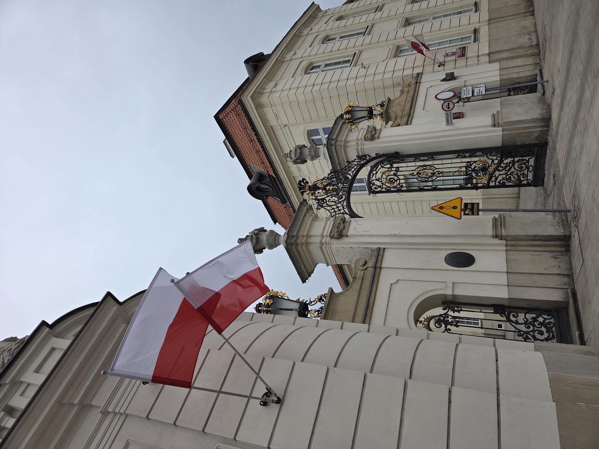 The image captures a view of the Royal Castle in Warsaw, Poland, taken at a low angle. In the foreground, two Polish flags are affixed to the building's facade, each displaying the national colors of white and red. The building is characterized by its classic architectural design, featuring light-colored stone or plaster walls, decorative moldings, arched windows, and ornate black wrought-iron gates. Above the entrance gate, there are intricate golden lampposts. Road signs are visible next to the gate. The sky is overcast.