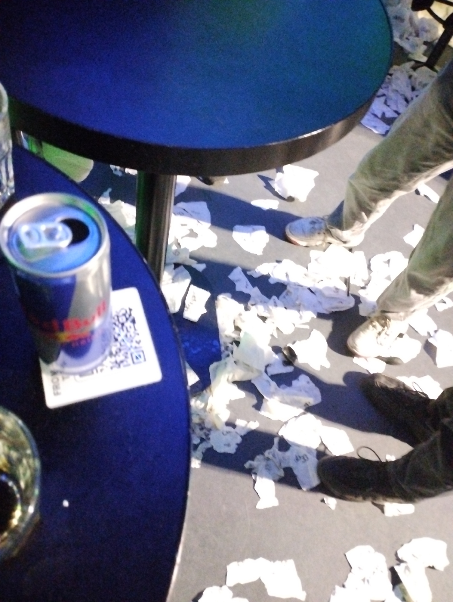 A Red Bull can sits on a coaster with a QR code on a blue table. There are two round, blue tables and paper scraps scattered on the floor. A person wearing jeans and light-colored sneakers and a person wearing black shoes are standing on the floor amidst the scattered scraps. The location is Unknown, Unknown.
