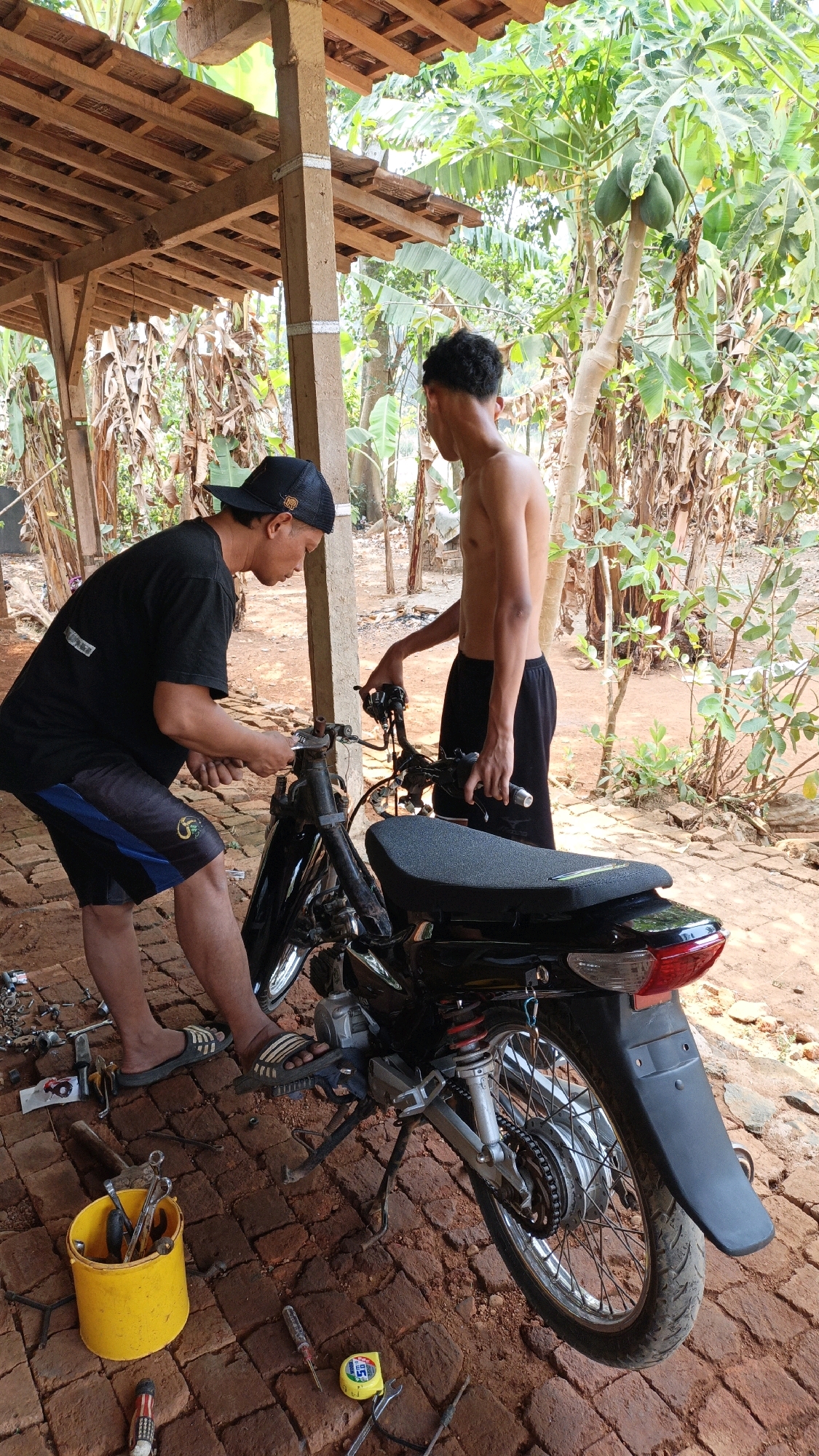 Two males are working on a black motorcycle under a wooden shelter in what appears to be a rural setting in Suwawal Timur, Indonesia. The person on the left, wearing a black t-shirt, black shorts with blue trim, and a black baseball cap, is holding a wrench and appears to be adjusting the handlebars or a related component. The person on the right, shirtless and wearing black shorts, is standing and observing, with one hand on the motorcycle's handlebars. The motorcycle is partially disassembled, with various tools scattered on the red brick ground around it, including a yellow bucket filled with tools, screwdrivers, and a tape measure. Lush green foliage, including banana trees and what appear to be papaya trees with fruit, surrounds the area, suggesting an outdoor, possibly residential or workshop environment. The lighting indicates it is daytime.