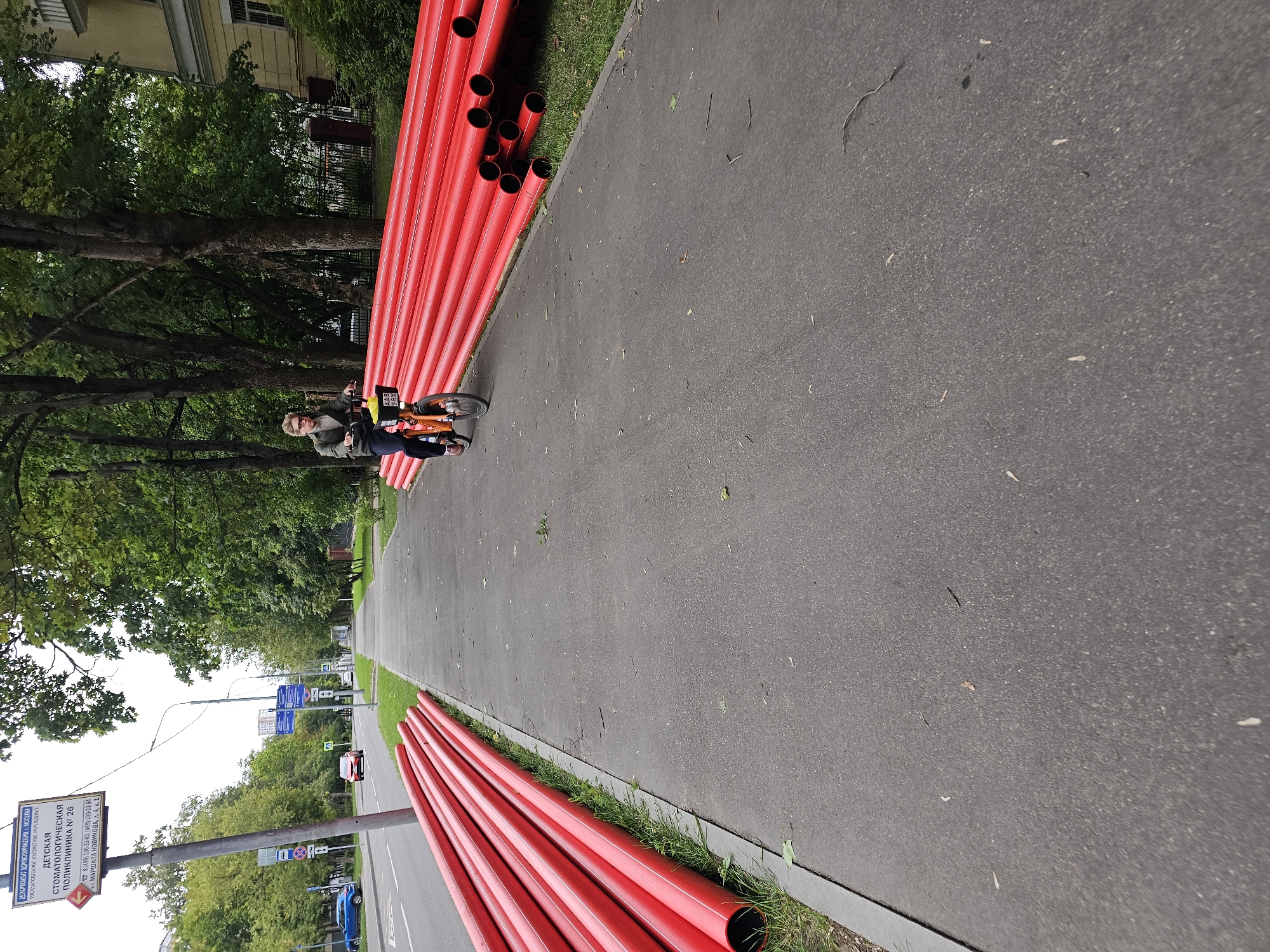 A person rides a bicycle along a paved sidewalk lined with a large stack of red, cylindrical pipes. The sidewalk is adjacent to a grassy area and a street with a few cars. In the background, trees and buildings are visible. A sign on a pole on the left side of the image appears to be in Russian. The image appears to be taken in a park or along a street in an urban environment, possibly in Khimki, Russia, given the presence of Russian signage.