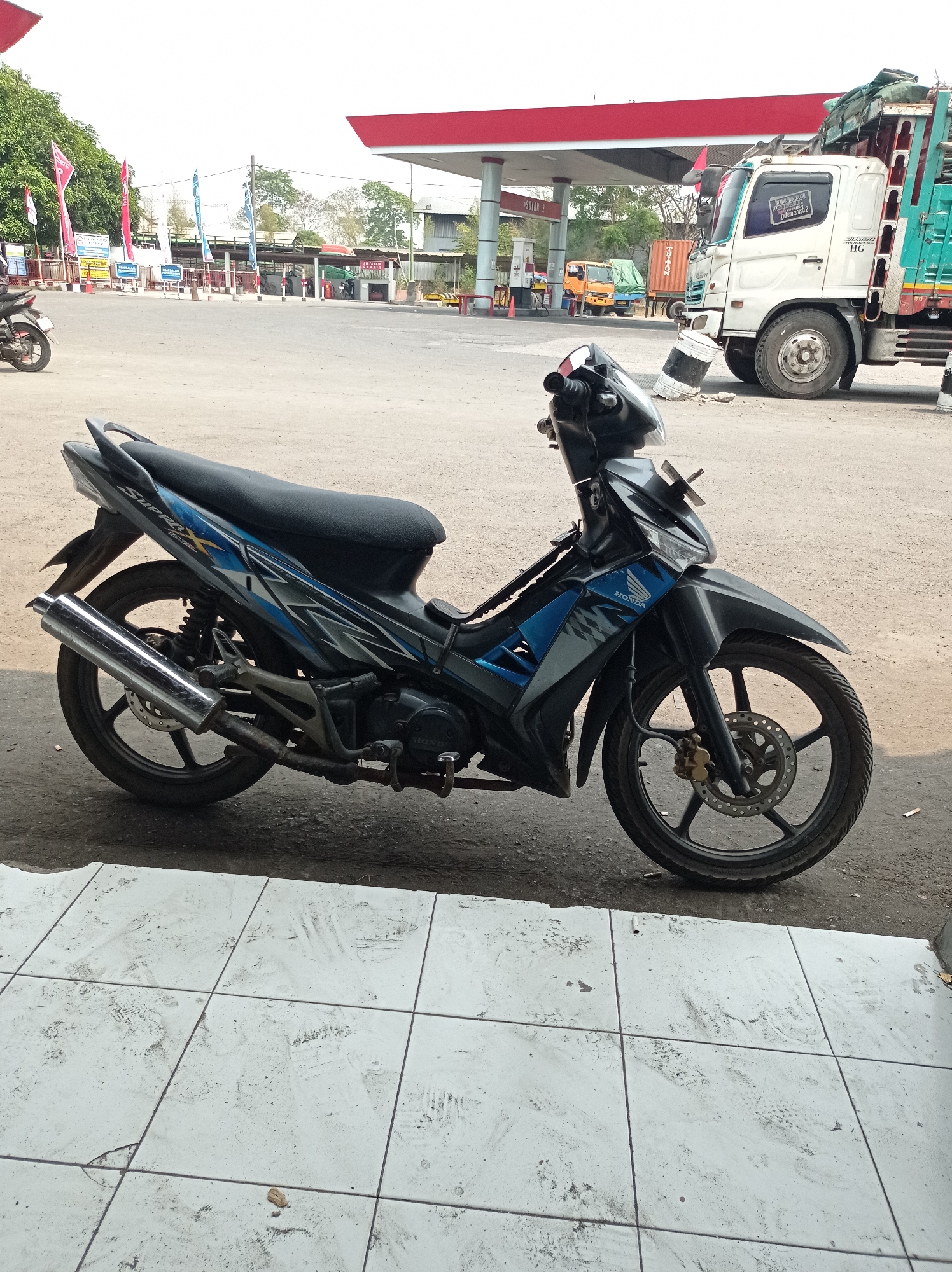 A black and blue Honda Supra X motorcycle is parked on a dusty, light-colored tiled surface, in front of a dirt and gravel lot. The motorcycle is facing to the right, with its rear wheel on the tiled surface and its front wheel on the dirt. The motorcycle has a chrome exhaust pipe and a black seat. In the background, a red-roofed gas station with a white truck and several other vehicles is visible. The gas station appears to be located in a city in Indonesia. The sky is clear and bright.