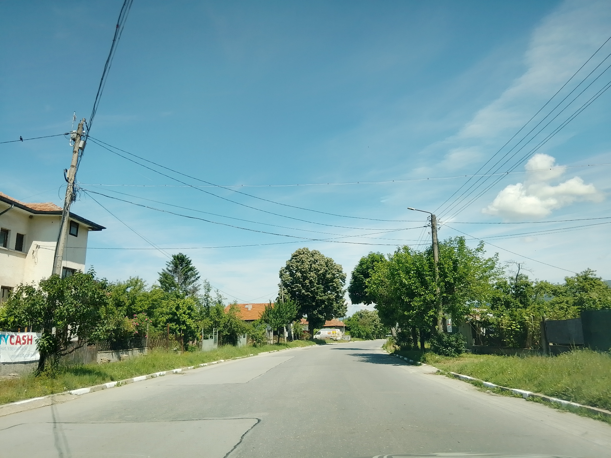 The image depicts a street scene in the city of Mirovo, Bulgaria, on a clear day with a bright blue sky and scattered clouds. The perspective is from a vehicle traveling along a paved road. On the left side of the road, a multi-story building with white walls and dark-framed windows is visible. Adjacent to the building is a banner with the text "CITYCASH". Further along the left side are several trees and a fence.
The road itself is gray asphalt with white and gray painted curbs. Overhead, a complex network of electrical wires and telephone lines crisscrosses the sky, supported by wooden utility poles.
On the right side of the road, there is a continuous line of lush green trees and vegetation. In the distance, more houses with red tile roofs can be seen amongst the trees. A white van with some visible markings is parked on the right side of the road in the mid-ground. The overall impression is a suburban or semi-rural setting.