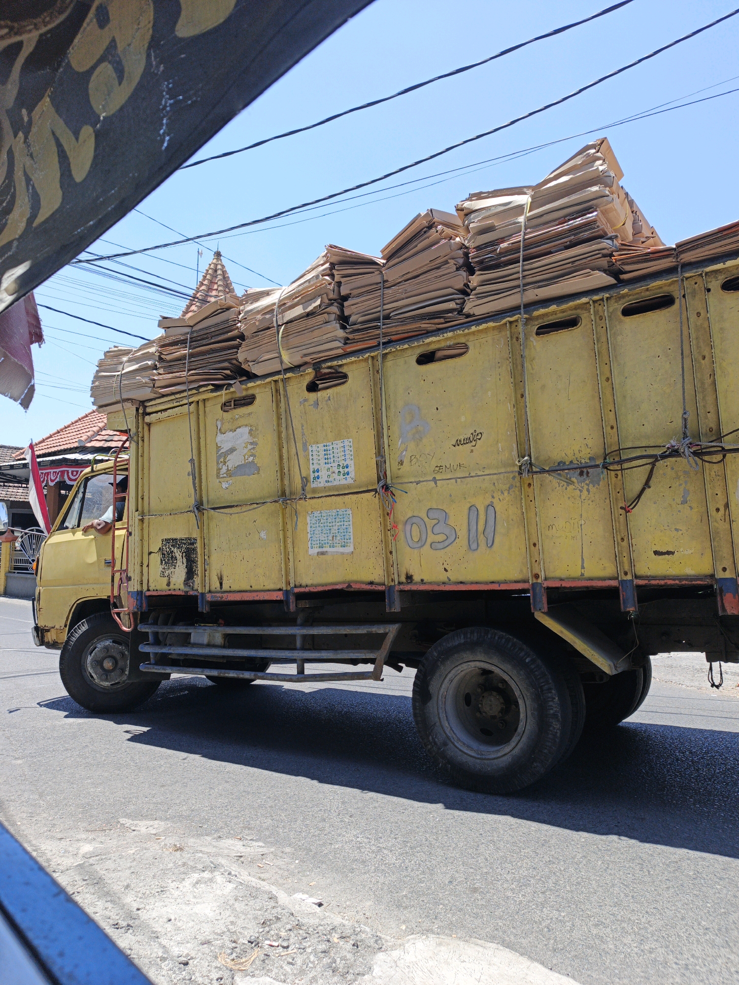 In Ketegan, Indonesia, a yellow truck loaded with a large stack of cardboard boxes is seen driving on an asphalt road under a clear blue sky. The truck has a vintage design, with visible rust and wear on its yellow paint. The cardboard boxes, tied down with ropes, extend high above the truck's bed, some leaning precariously. A red and white Indonesian flag is attached to a pole near the front of the truck, adding a touch of national identity. The background reveals traditional Indonesian houses with tiled roofs. The image captures a moment of daily life and commerce in this Indonesian town, with the truck likely transporting goods to or from a business.
