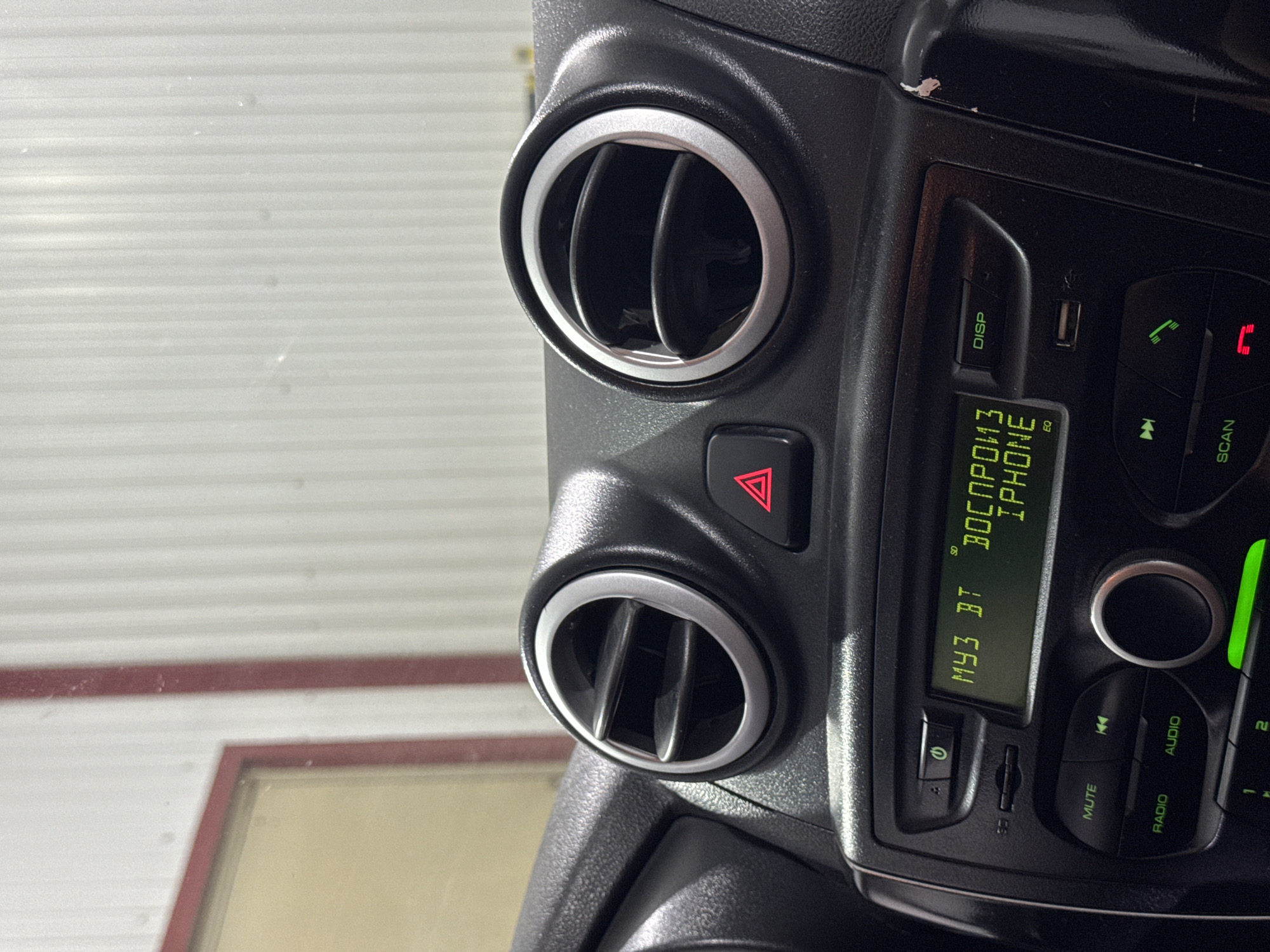 The image shows the interior of a car, specifically the dashboard area. In the center, a car stereo system is visible with a green LCD display. The display shows the text "МУЗ BT" which likely indicates music playing via Bluetooth. To the right of this, it reads "ВОСПРОИЗ" and "PHONE", suggesting "playing from phone". There are also air vents on either side of the stereo, and a hazard light button with a red triangle symbol is located between the vents. The dashboard is black plastic and shows some wear and tear, particularly on the right side. The background of the image is blurred but appears to be a white, corrugated surface, possibly a wall in a garage or workshop. The image was taken in Илсхан-Юрт, Russia.