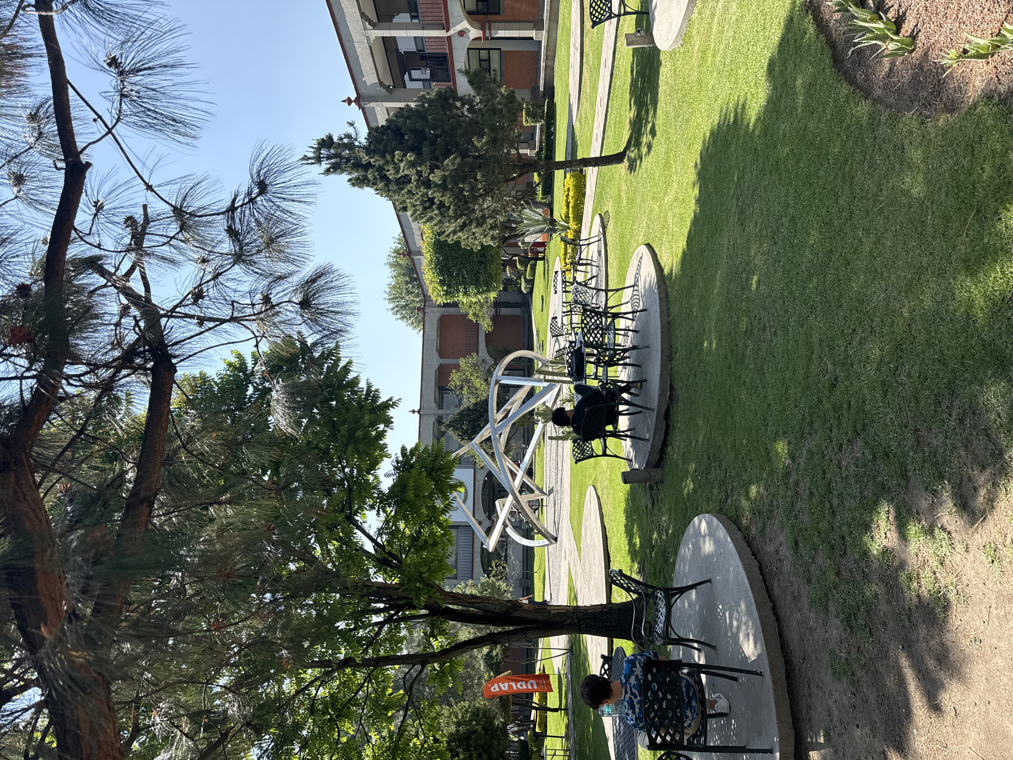 The image depicts a courtyard or garden setting in San Andrés Cholula, Mexico. The scene features a well-maintained lawn, trees providing shade, and a pathway leading through the green space. Seating areas with tables and chairs are scattered throughout the courtyard, some occupied by individuals. In the background, a multi-story building with architectural details is visible. A modern, abstract sculpture made of metal stands prominently in the garden. An orange rectangular sign with the letters "UDLAP" on it is visible behind a tree. The weather appears to be sunny, casting shadows from the trees and building.