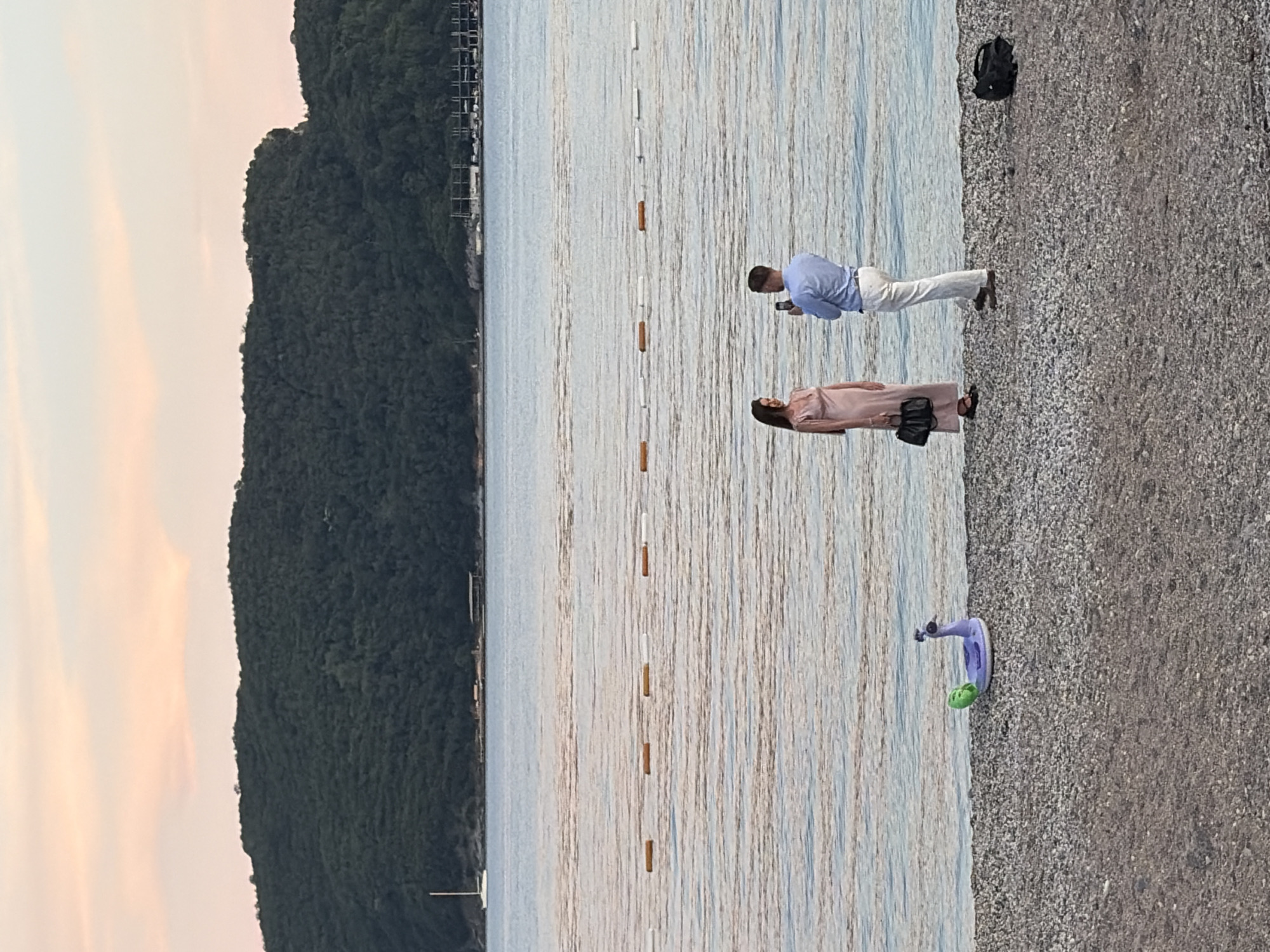 The image depicts a beach scene at dusk in Budva, Montenegro, with a body of water in the background and a forested hill beyond that. In the foreground, a pebble beach is visible. A purple inflatable peacock with a green accent is lying on the left side of the beach. Two people, a woman in a long, light pink dress and a man in a light blue shirt and white pants, are standing near the water's edge. The man is holding a phone and appears to be taking a photo of the woman, who is looking towards him. A black handbag is resting on the ground to the right of the man, and another black bag is further to the right. The water is calm, with a visible line of buoys separating the swimming area. The sky shows pastel colors of pink and orange, indicating sunset. A white sailboat is visible in the distance on the left, and structures resembling docks or buildings are present along the shoreline of the hill.