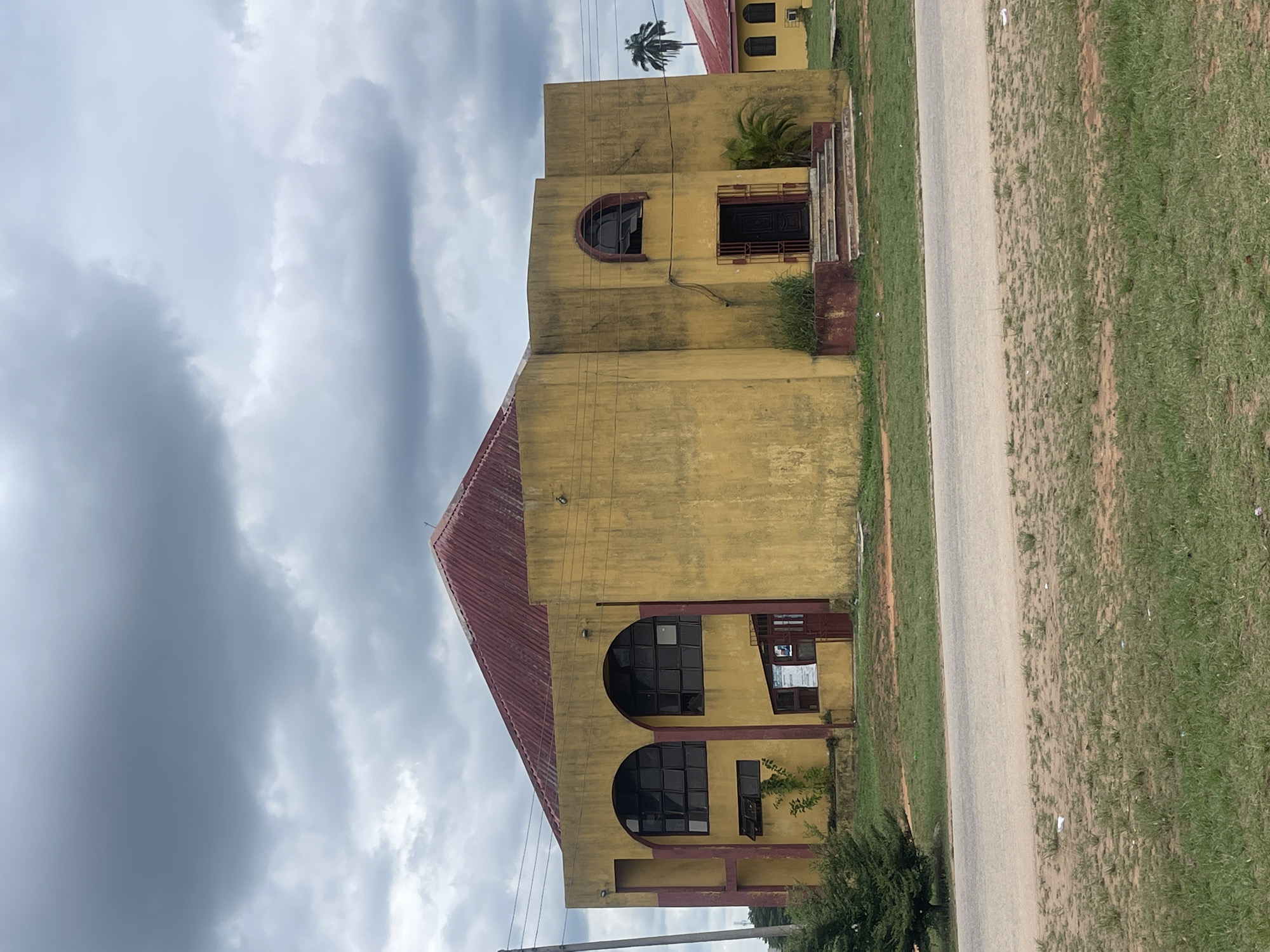 The image shows a two-story yellow building with a red roof in Abraka, Nigeria. The building is located next to a paved road with grass on both sides. The sky is cloudy. The building features arched windows and doors on both floors. A palm tree is visible in the background.
