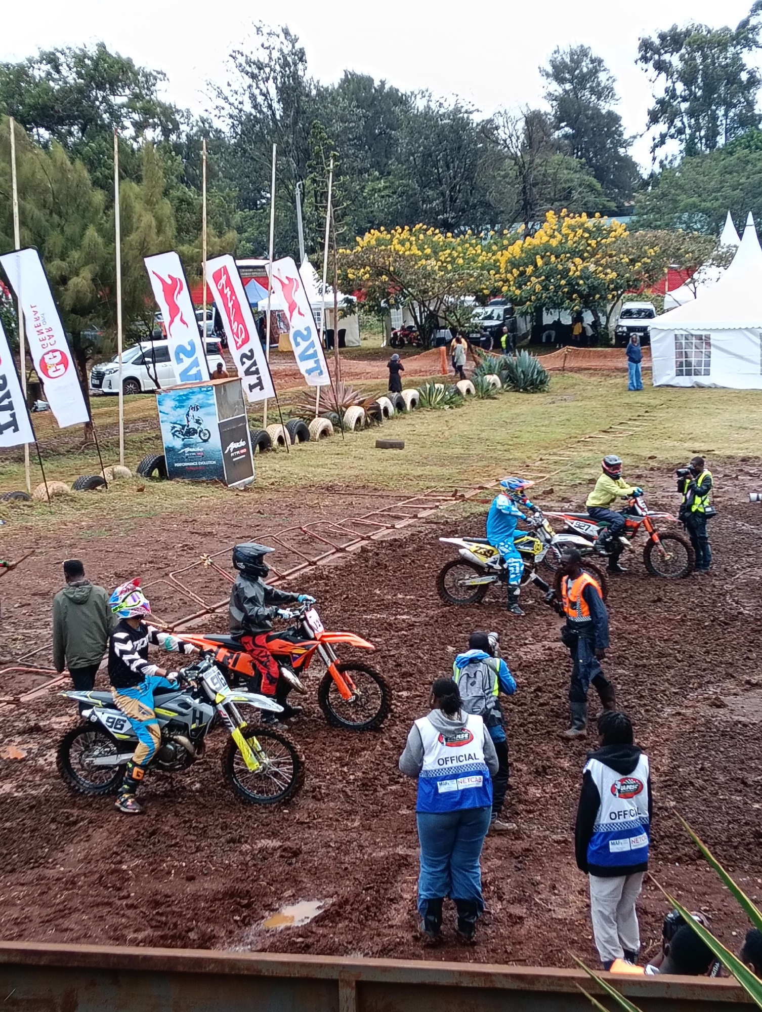 Here's a detailed description of the image:
**Scene Overview:** The photo captures a motocross event taking place on a muddy course. It appears to be an amateur or semi-professional event, with participants and spectators present. There are tents, flags, and vehicles indicating a structured event space.
**People:**
* Multiple motocross riders are on their bikes, some in racing attire. They're wearing helmets and protective gear. Numbers are visible on their bikes (e.g., 96, 927, 259).
* Several individuals are wearing "Official" vests, indicating they are event staff.
* Spectators and other individuals are present, some dressed casually. A person is seen filming with a camera.
**Subjects/Objects:**
* Motocross bikes are prominent.
* Flags bearing the logos of "2VT" are visible.
* A sign with motocross-related graphics is present.
* Vehicles are parked in the background.
* Tents are set up in the background.
* The ground is very muddy, showing signs of recent rain or track watering.
**Actions/Interactions:**
* Riders are maneuvering their bikes on the muddy course, seemingly preparing to race.
* Officials are observing and likely directing the participants.
* Spectators are watching the event.
**Environment:**
* The location seems to be an open field or park area on the outskirts of Nairobi, Kenya.
* Trees and other greenery surround the area, hinting at a semi-rural location. The sky is overcast.
**Location Details:** Due to specific details like license plates or precise landmarks being absent, a pinpoint location cannot be forensically determined. However, it can be inferred that the scene is within or near Nairobi, Kenya.