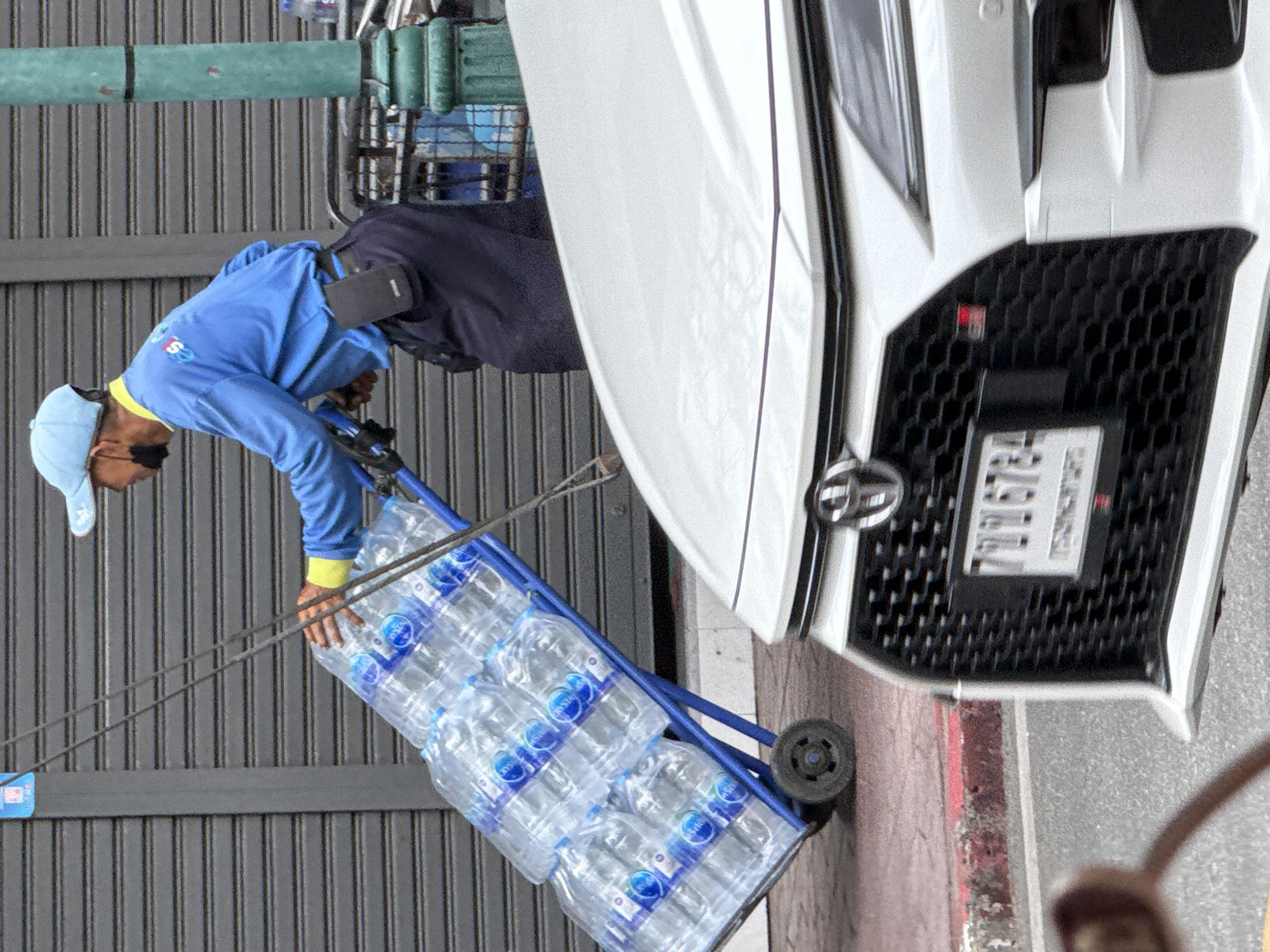A person wearing a blue shirt, blue cap, and dark pants is pushing a blue cart laden with cases of bottled water. The cart has two wheels. The person is wearing a black face mask. A white car with a prominent grille and a license plate is in the foreground. The scene appears to be on a street in Bangkok, Thailand, indicated by the street signs and architecture. The background features a gray, corrugated metal shutter.