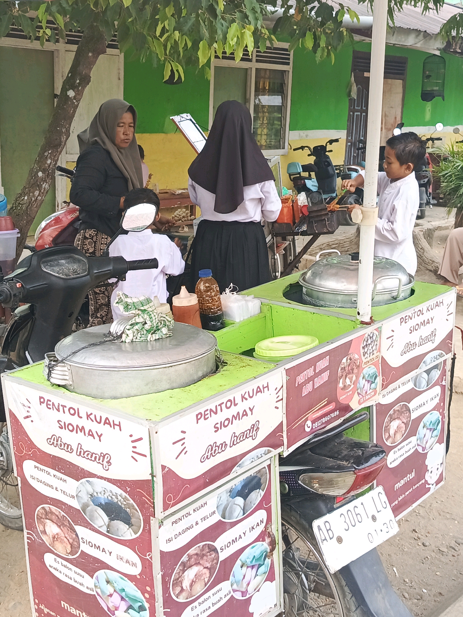 The image depicts a street food vendor in Megang Sakti II, Indonesia. The vendor, a woman wearing a hijab and a dark jacket, is seated on a red motorcycle. Beside her, a young boy in a white shirt and pants is preparing food at a mobile cart. The cart is green and adorned with colorful advertisements for "Pentol Kuah Siomay Abu Hanif," showcasing images of food items like siomay, meatballs, and drinks. The background reveals a building with green and yellow walls, suggesting an outdoor commercial area. The overall scene is bustling, with signs of daily commerce and community interaction.