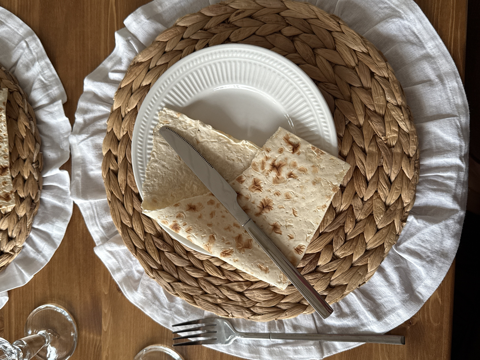 The image displays a close-up, overhead view of a place setting on a wooden table. The setting includes a woven placemat with a ruffled white cloth underneath. Centered on the placemat is a white plate with a ribbed pattern. On the plate are two pieces of matzah, one folded over the other, with a silver knife resting on top. To the left of the placemat, a silver fork is visible. In the upper left corner, a portion of a clear glass with a stem is present. The overall scene depicts a prepared meal or a dining arrangement. The location is identified as Starokorsunskaya, Russia.
