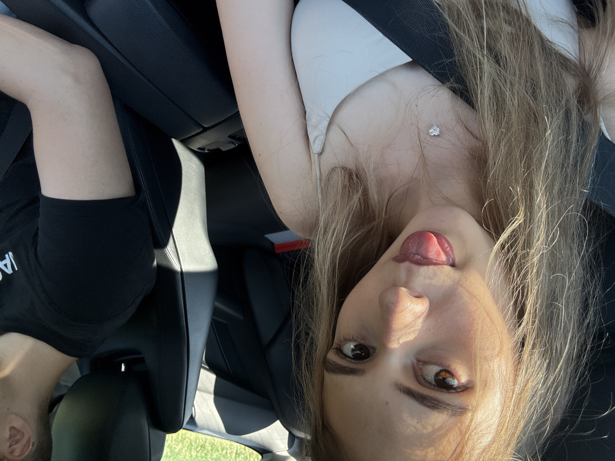 The image shows two people in a car, with the camera angled downwards. The person in the foreground, on the right, is a woman with long blonde hair. She is wearing a white top and has her tongue sticking out. She is also wearing a necklace with a small diamond pendant. The person in the background, on the left, is a man wearing a black t-shirt. He appears to be the driver of the car. The car's interior is visible, including the black leather seats and a seatbelt. The scene appears to be set during the day, with sunlight coming through the windows. The background outside the car shows greenery, suggesting a rural or suburban location. The overall mood of the image is playful and casual. The visible text on the man's t-shirt is "IA". The location context is Iffezheim, Germany.