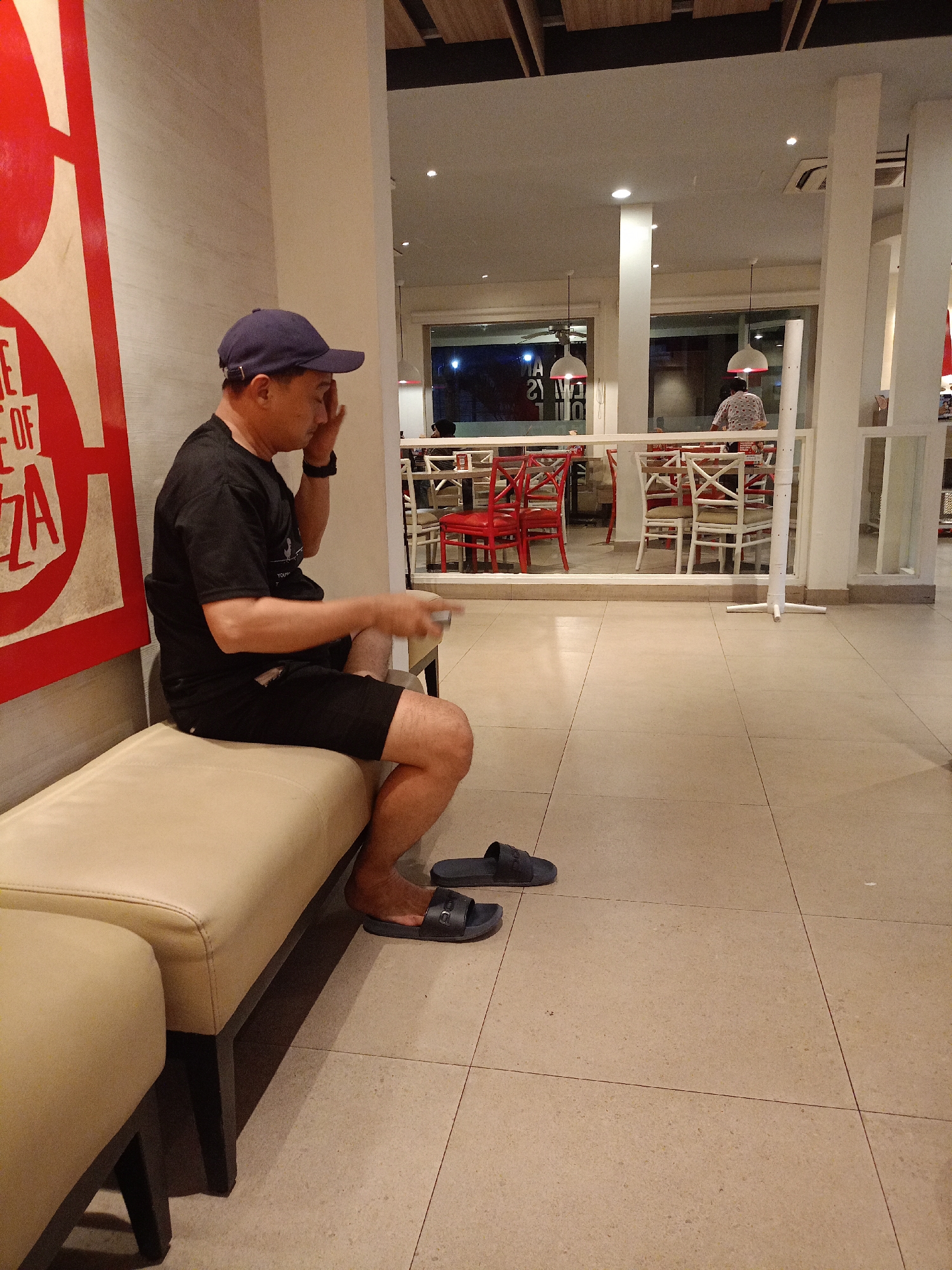 A man wearing a dark blue baseball cap, a black t-shirt, and black shorts is sitting on a light-colored bench. He is holding a small white object in his right hand and appears to be touching his forehead with his left hand. He is wearing dark blue slide sandals. The man is positioned on the left side of the frame, facing towards the right.
Behind the man, the scene depicts the interior of a restaurant, likely a Pizza Hut, given the prominent red and white signage with the word "PIZZA" visible on the left. There are tables and chairs in the background, with some chairs being red and others white. The restaurant appears to be well-lit with ceiling lights and pendant lamps. The floor is tiled in a light beige color.
The location is the Pizza Hut restaurant on Jalan Sultan Agung, Batu, Indonesia.
