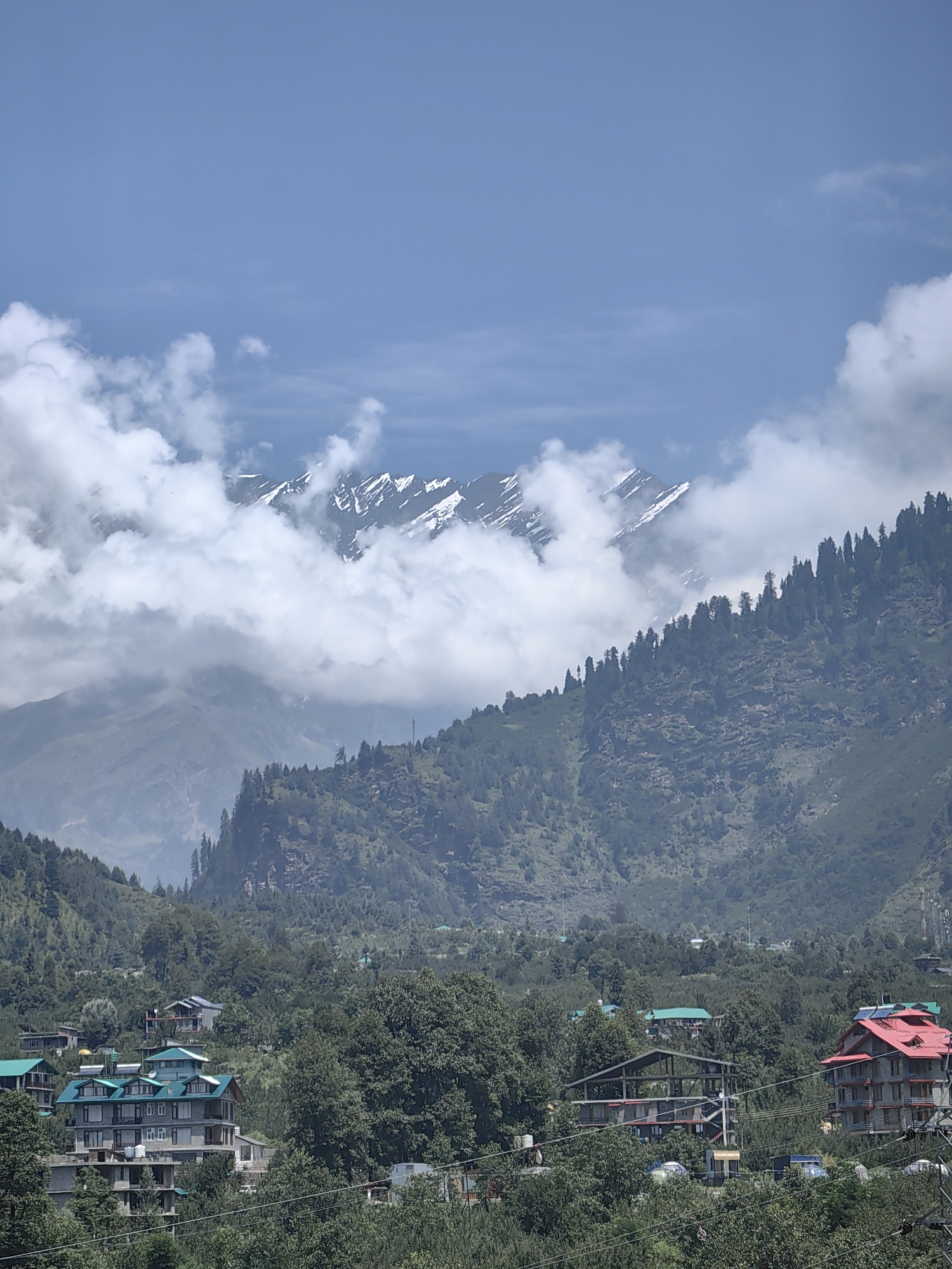 This is a photograph taken from a high vantage point, looking towards a mountain range covered in snow and clouds. In the foreground, a lush green valley is dotted with buildings and trees. The sky is a clear blue with a few scattered white clouds. The scene appears to be a scenic landscape in the city of Kullu, India, possibly in the area of Jagatsukh or Vashisht, given the presence of buildings with tiered roofs and stone facades often seen in the region. The image captures a serene mountain vista with a blend of natural beauty and human settlement.