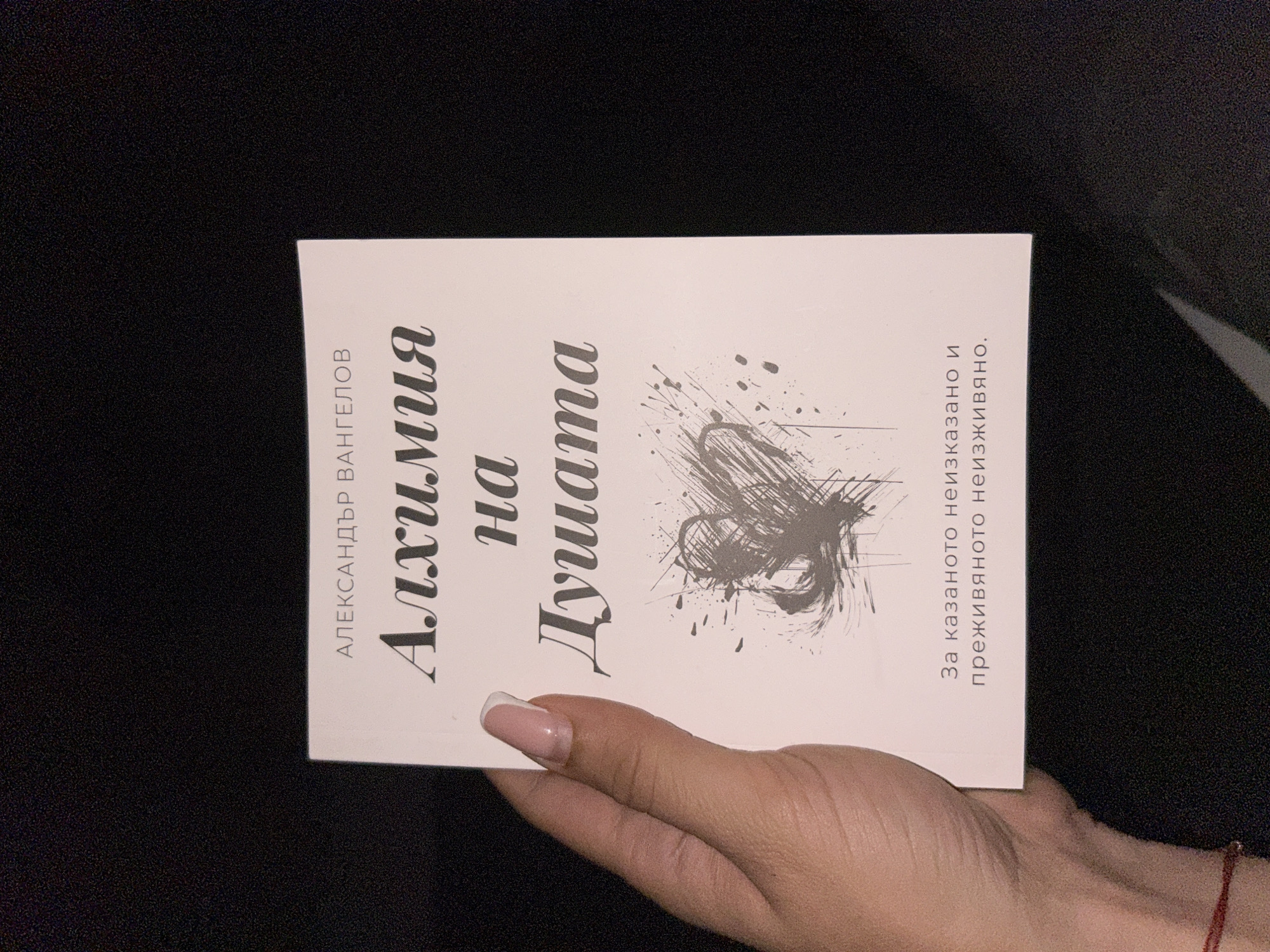 A person's hand with manicured nails is holding a book. The book has a pale pink cover with text in a dark color. The title of the book appears to be "Алхимия на душата" (Alchemy of the Soul). Below the title, there is a graphic design resembling a heart with splatters. The bottom of the book has additional text. The background is dark and appears to be out of focus. The lighting suggests it is indoors. The location is unknown.