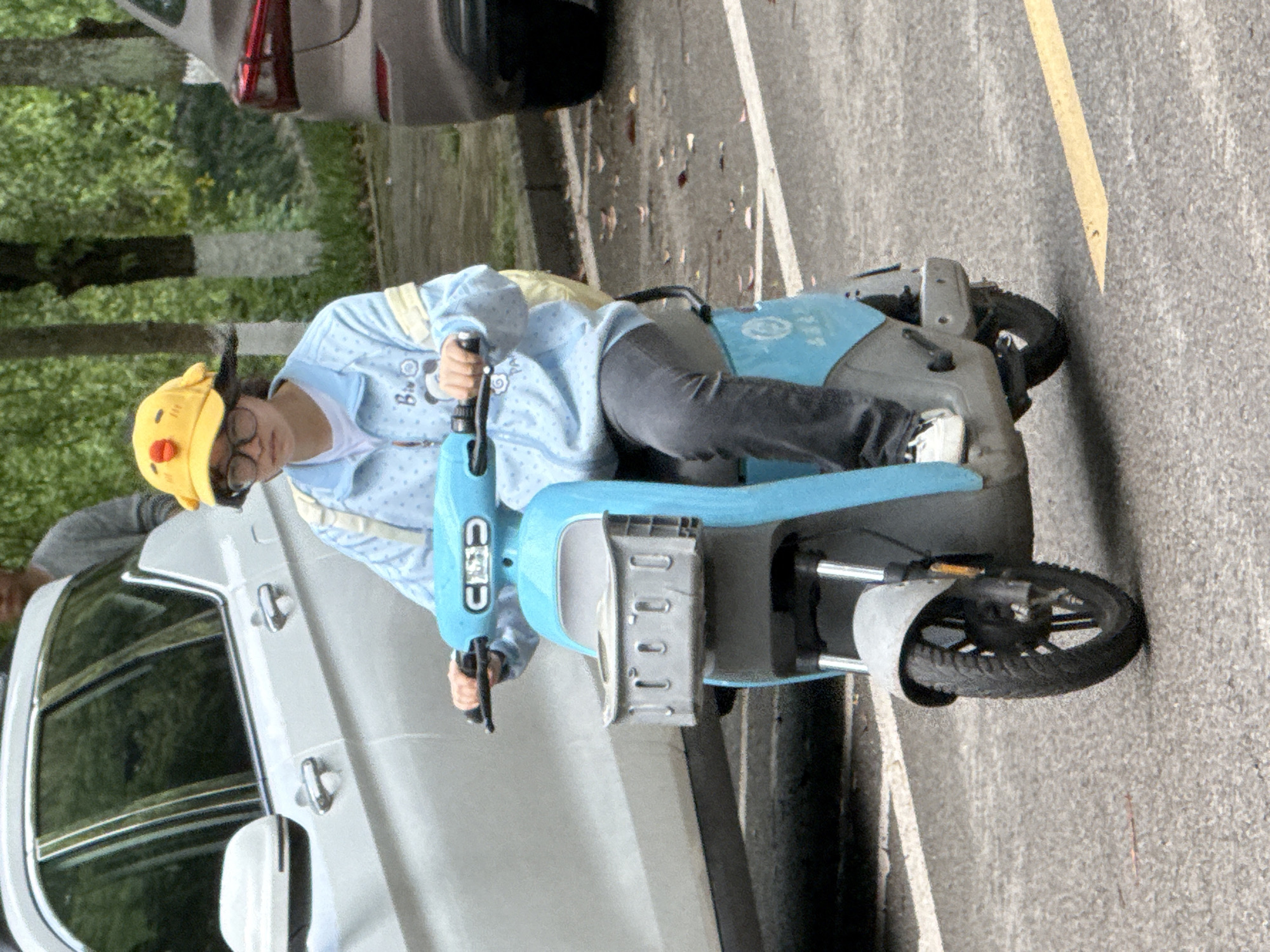 A person wearing a yellow chicken hat, glasses, and a light blue patterned jacket is riding a light blue and gray electric scooter on an asphalt road next to parked cars. The location is in Jiangjin, China. A yellow line marking is visible on the road. Trees are visible in the background.