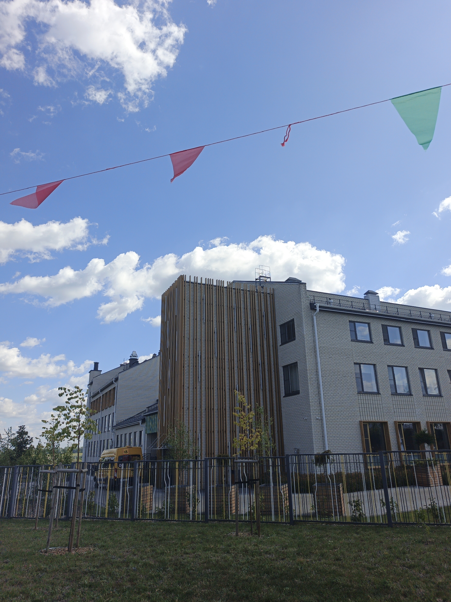The image shows a modern, light-colored brick building with numerous windows, possibly a school or administrative facility, with a decorative wooden facade element. The building is set against a bright blue sky with scattered white clouds. A string of triangular pennants, some pink and one green, is stretched across the top of the image. In the foreground, there is a black metal fence with vertical bars of various colors, including yellow and white. Beyond the fence, a grassy area with young trees is visible, along with a yellow bus. The environment appears to be an outdoor urban setting, possibly a park or school grounds, in the city of Repnoe, Russia. There are no people visible in the image.