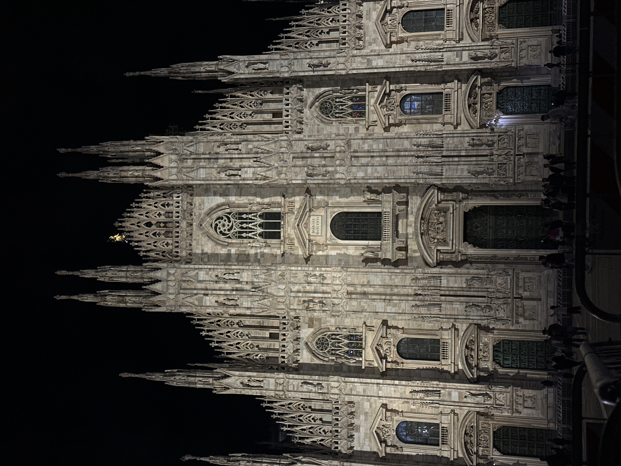 The image shows the Milan Cathedral (Duomo di Milano) at night, illuminated against a dark sky. The facade exhibits intricate Gothic architectural details, including spires, rose windows, sculptures, and decorative carvings. Several people are present on the right side of the frame, near the base of the cathedral, viewed from a low vantage point. A metal railing is partially visible in the foreground on the right.