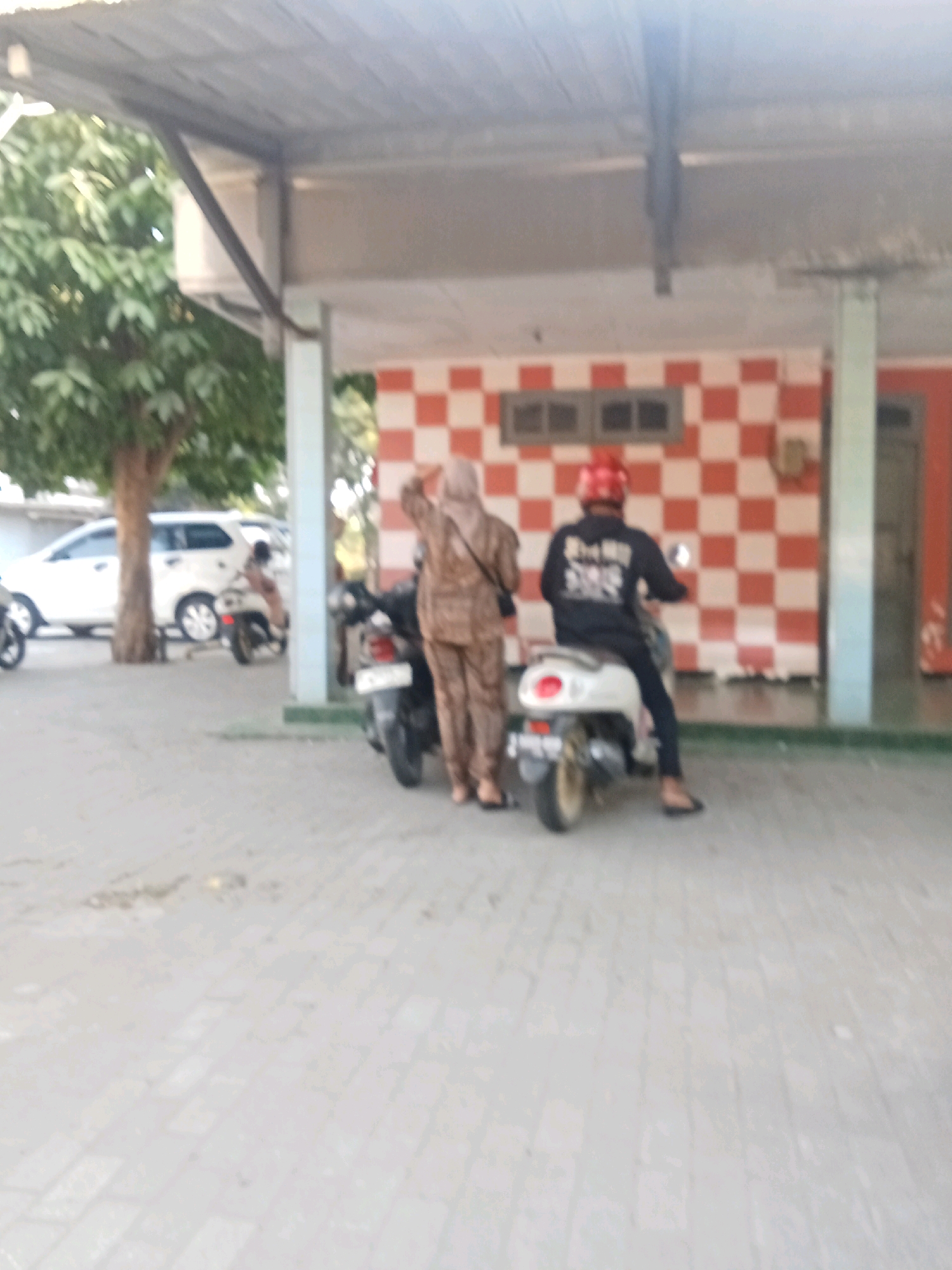 The image depicts an outdoor scene in what appears to be a covered walkway or a parking area in Kaliwungu, Indonesia. The foreground is paved with light-colored rectangular stones. In the midground, two people are visible on motorbikes. On the left, a person wearing a patterned outfit and a headscarf is standing next to a parked motorbike. On the right, a person wearing a black long-sleeved shirt and a red helmet is seated on a white motorbike. Behind them, a building features a wall with a red and white checkered pattern and a small, rectangular window. A white SUV is parked on the left side of the image, partially obscured by a tree. The roof structure above is made of corrugated metal supported by beams. The lighting suggests it is daytime, with sunlight creating shadows and highlights.