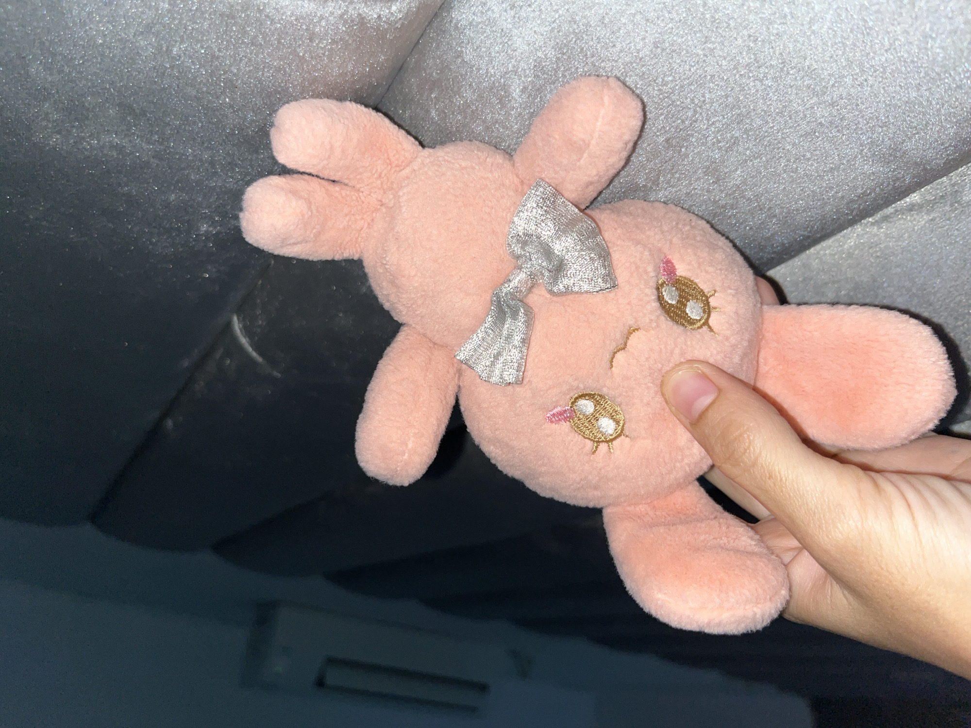 The image features a close-up shot of a human hand holding a pink plush toy indoors. The plush toy, which resembles a rabbit or similar animal, is light pink and has a soft, fuzzy texture. It is adorned with a silver, shimmery ribbon tied in a bow on its head. Its face has embroidered features: two large, golden-brown eyes with white highlights and black eyelashes, and a small pink/red detail that could be a nose or mouth. The toy has four limb-like appendages and two longer, ear-like protrusions.
A person's hand, with visible skin folds and fingernails, is holding the lower body of the plush toy, positioning it prominently in the frame.
The background is divided into two main sections. The upper portion is a light gray or silver surface with a noticeable texture, possibly a fabric like a cushion or upholstery, or a textured wall/ceiling. This area shows some light reflections, suggesting artificial lighting. The lower background is dark, appearing black or very dark blue, and has a somewhat reflective quality. Faint dark shapes or objects are visible in this darker area, hinting at furniture or other room elements, and possibly part of an air conditioning unit can be discerned in the bottom left. The lighting appears artificial, suggesting the photo was taken indoors, possibly at night or in a dimly lit environment. No text is visible in the image. The scene conveys a casual, personal moment.