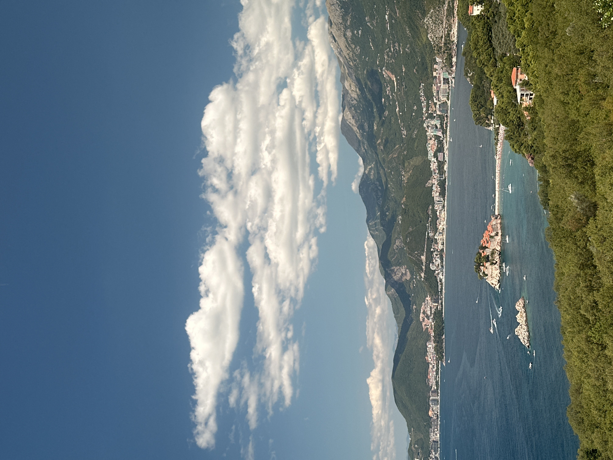 A high-angle, slightly over-the-shoulder view captures the Adriatic Sea and the coastal town of Sveti Stefan, as seen from Blizikuće, Montenegro. The deep blue water is dotted with several small boats and yachts, some creating wakes on the surface. In the foreground, lush green trees dominate the landscape, with glimpses of red-tiled roofs of houses peeking through the foliage.
The focal point of the image is the iconic island of Sveti Stefan, connected to the mainland by a narrow causeway. The island is densely packed with stone buildings, appearing as a cluster of ancient structures. A beach is visible on the mainland near the causeway, with umbrellas and people enjoying the sun.
Further in the background, a long stretch of coastline unfolds, featuring a modern city with numerous buildings, hotels, and resorts. These structures are nestled against the backdrop of rolling green hills that gradually rise into rugged, rocky mountains under a bright blue sky. Scattered white clouds add texture and depth to the sky. The overall scene depicts a scenic coastal panorama, characterized by the contrast between the natural beauty of the mountains and sea, and the developed urban and resort areas.