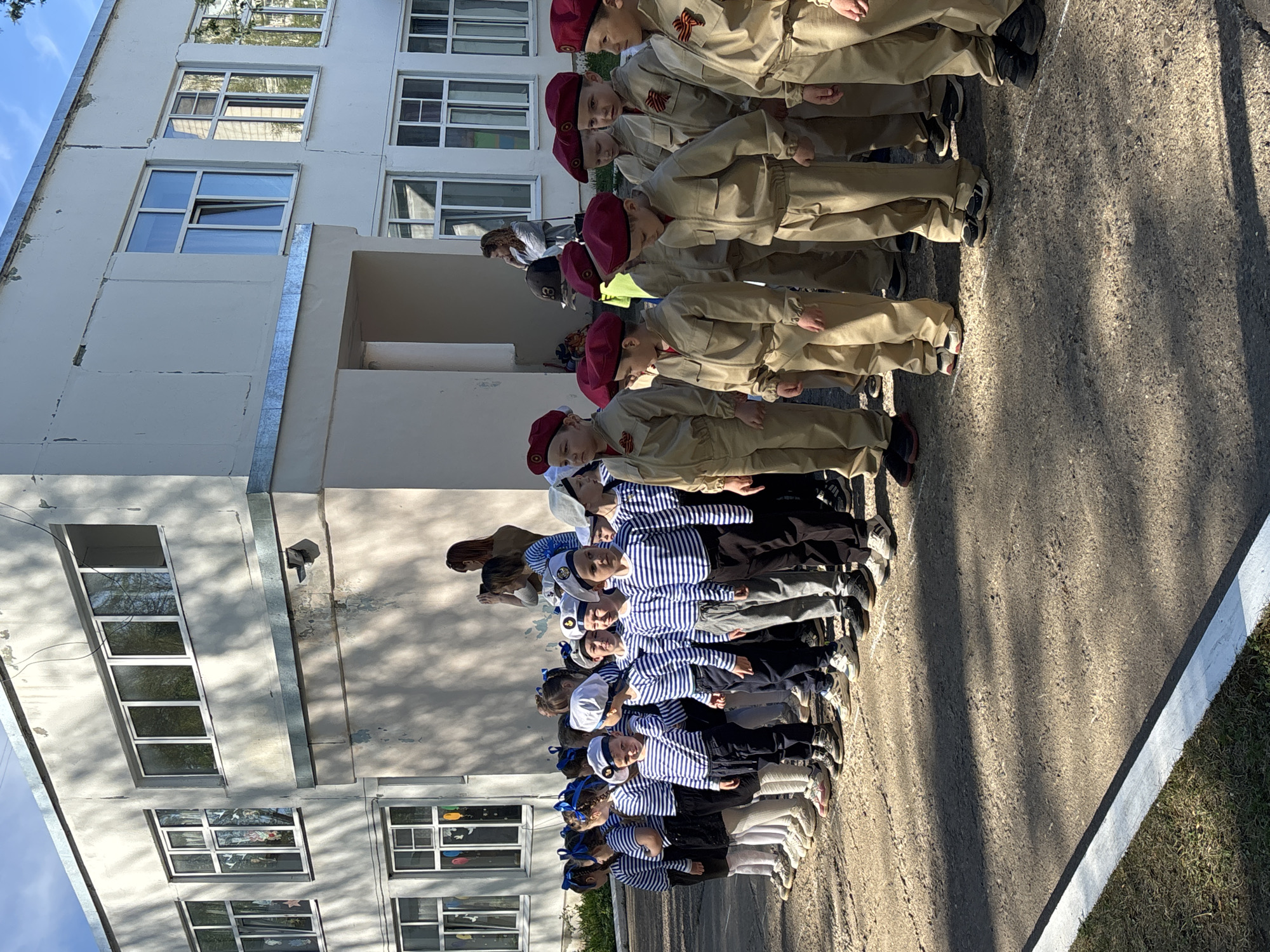 In the city of Chaikovsky, Russia, a group of children are lined up outside a multi-story, light-colored building. Some of the children are wearing sailor uniforms with blue and white striped shirts and sailor hats. Others are wearing tan uniforms with red berets and red star pins. They appear to be standing at attention. The location is outdoors on a paved surface next to a building. The weather is sunny, casting shadows on the ground.
