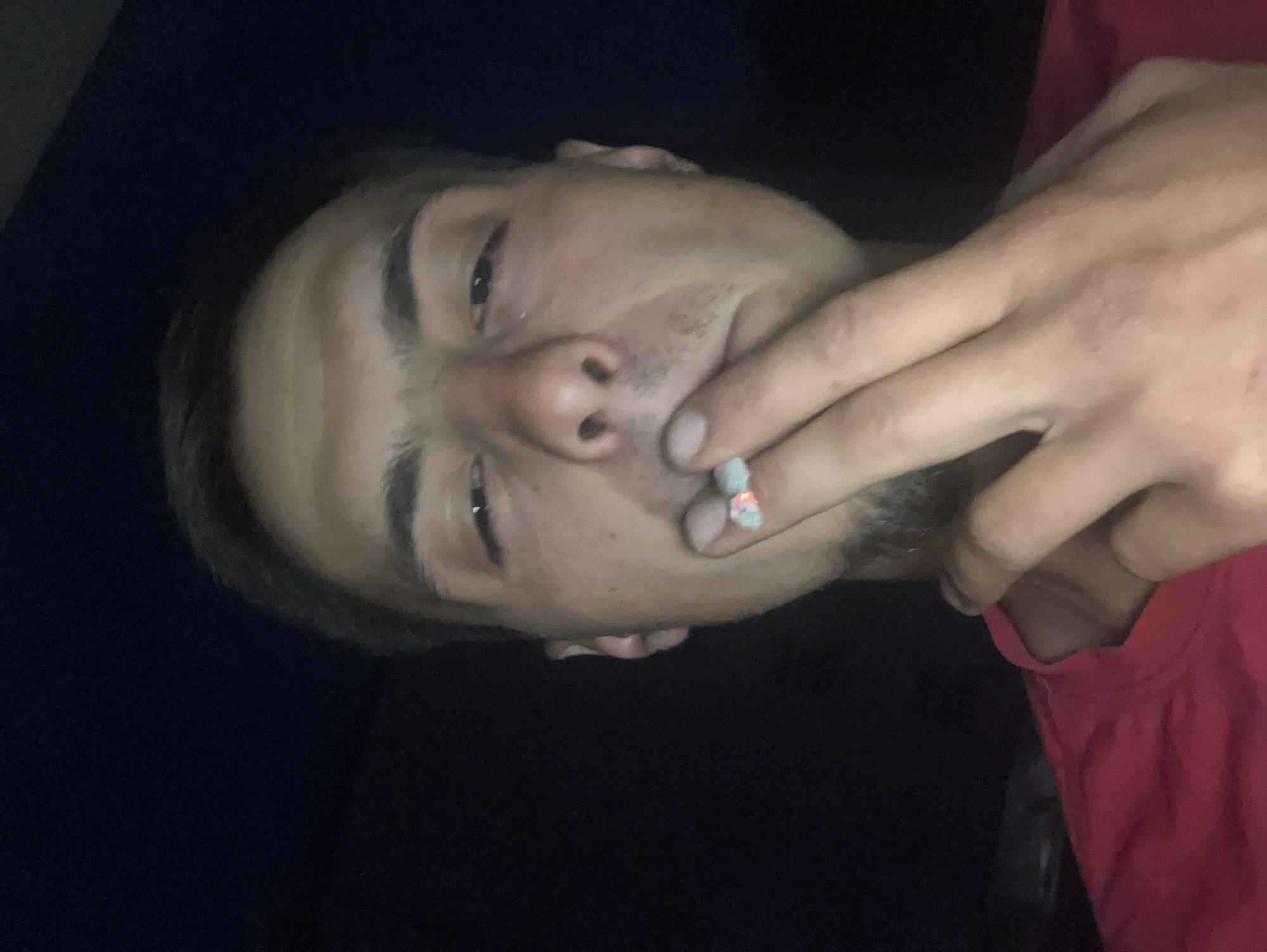 The image depicts a man holding a lit cigarette between his fingers, close to his mouth. He is facing the camera with a slight downward gaze. The background is dark, with a portion of a red garment visible on the right. The image does not provide any information to determine the location in the city of Plovdiv, Bulgaria.