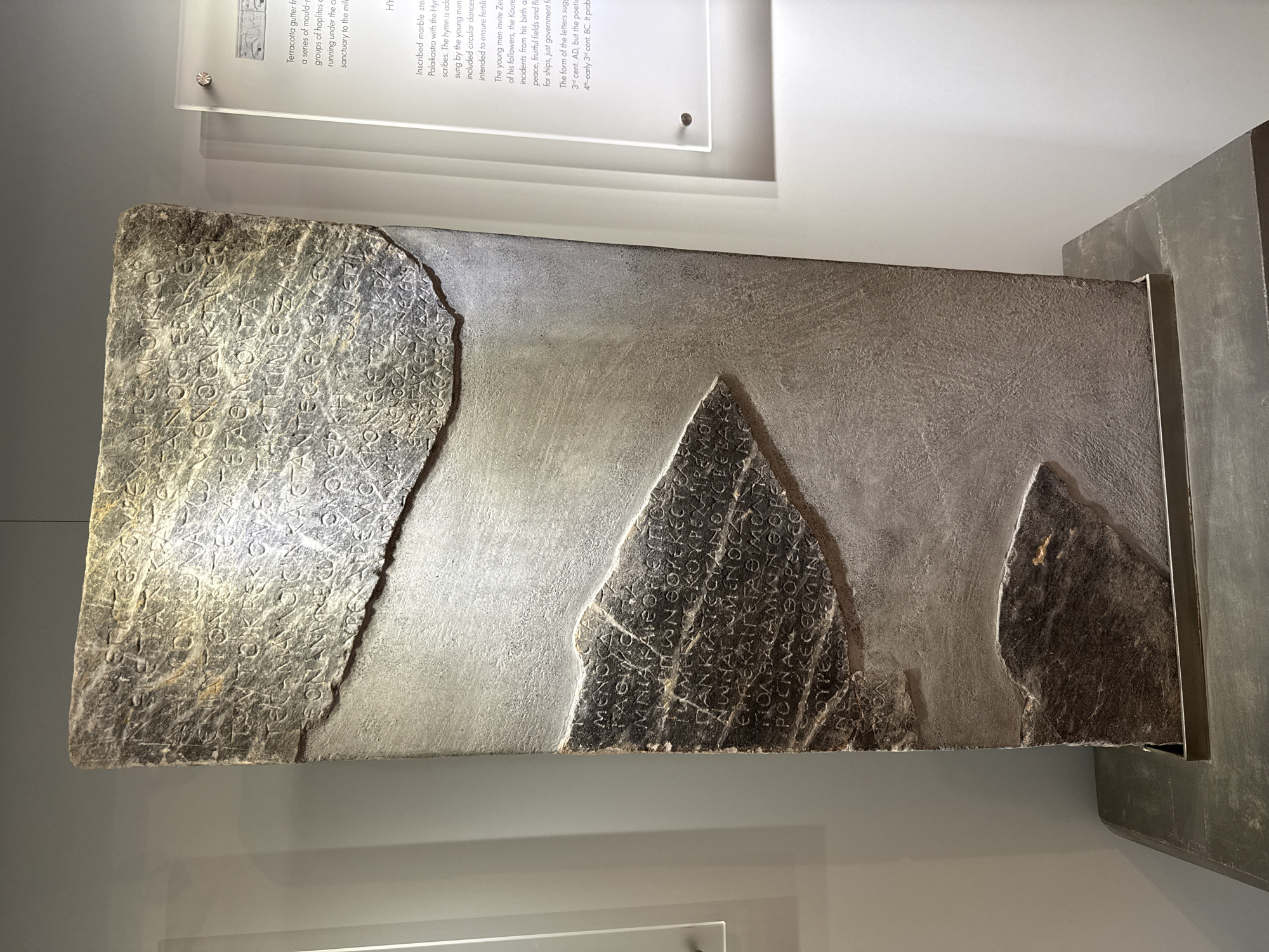 The image shows a large, rectangular stone stele with inscriptions. The stele is dark gray and appears to be made of marble or a similar stone. It is displayed vertically on a stand in what appears to be a museum setting. There are two main sections of inscriptions visible, with a large piece missing from the upper right and a smaller piece missing from the lower right. The text is carved into the stone in an ancient script.
To the right of the stele, a small information panel is visible, made of clear acrylic with text printed on it. The panel has a title that reads "Hymn". The text on the panel describes the stele as an "Inscribed marble stele from Palaikastro with the Hymn to Zeus". It mentions that the hymn was sung by young men, included circular dances, and was intended to ensure fertility. It also notes that the young men invite Zeus and his followers to join them, and the hymn is connected to events from Zeus's birth and subsequent life. The panel also mentions the form of the letters suggesting a date of "4th-early 3rd cent. BC".
While the city of Heraklion, Greece, is mentioned in the prompt as a potential location, this information cannot be visually confirmed from the image itself. The environment appears to be an indoor exhibition space with neutral-colored walls and subtle lighting. No people are visible in the image.