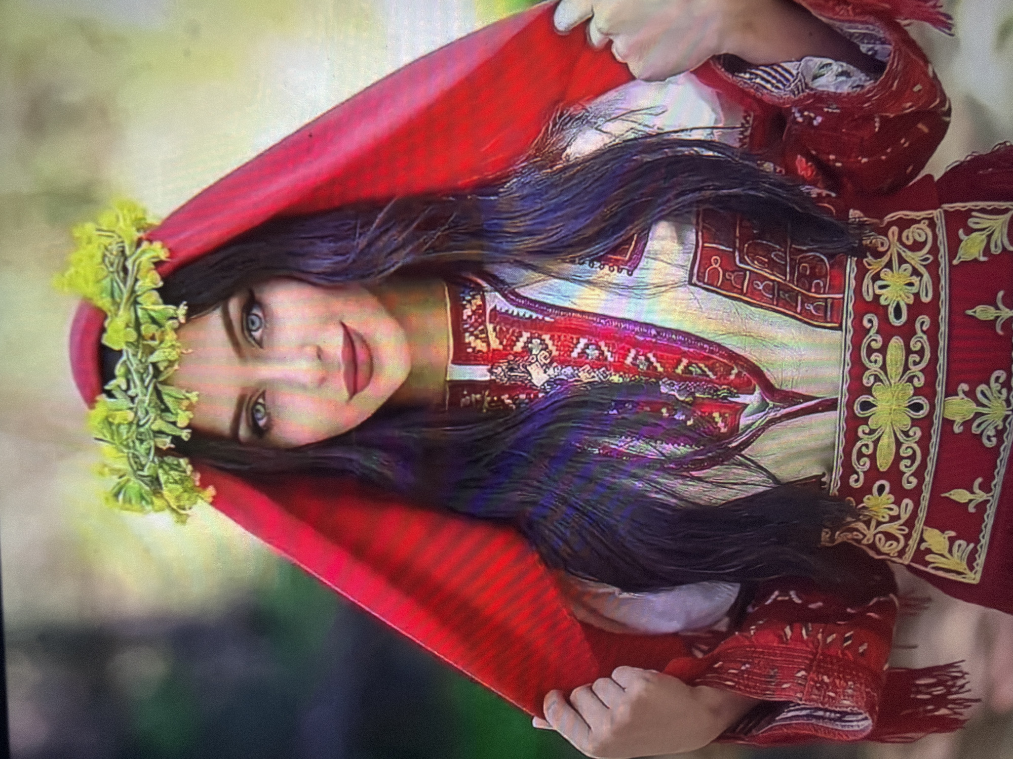 A female subject is centrally positioned, facing forward with a direct gaze and a subtle closed-mouth smile. She possesses dark, wavy hair, light blue eyes enhanced with makeup, and deep red lipstick. Her head is enveloped by a bright red fabric, draped to frame her face and shoulders, topped with a wreath composed of green foliage or small yellowish-green blossoms. She is attired in what appears to be traditional clothing: a light-colored tunic or dress bodice meticulously embroidered with red and white geometric and floral patterns. The sleeves exhibit similar red and white embroidery, potentially with fringed cuffs. A wide red belt, richly decorated with gold-colored scrollwork or floral motifs, encircles her waist. Both hands are visible, positioned at either side of her face, lightly holding the red head covering. The background is blurred, consisting of diffused green and yellow tones, indicative of an outdoor, natural environment such as foliage. The scene is stated to be located in the city of Shumen, Bulgaria, though no discernible urban architectural elements are present within the visual frame to corroborate this specific location.