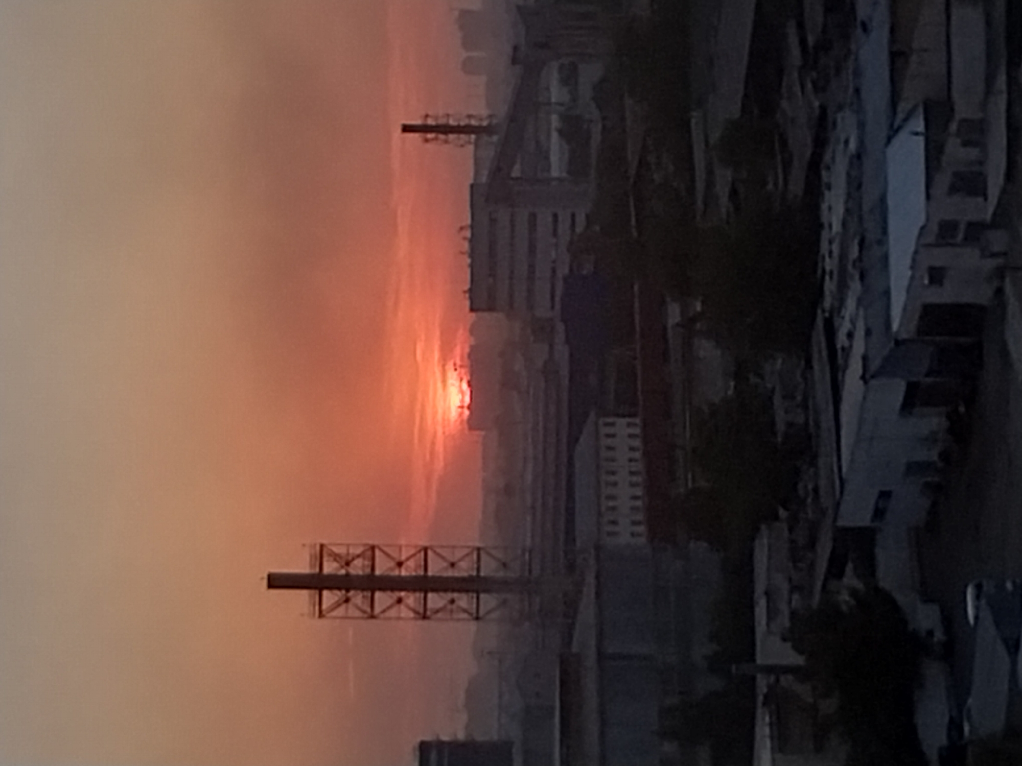 The image depicts an industrial area in Tashkent, Uzbekistan, during a sunset. The sky is a hazy orange and red, indicating atmospheric pollution or dust. In the foreground, several low-rise industrial buildings are visible, with some featuring corrugated roofs. Further in the background, a tall, lattice-like industrial tower stands prominently, silhouetted against the sunset. To the right of this tower, another, more solid industrial structure with a vertical chimney rises. In the distance, larger, more modern buildings and possibly apartment complexes can be seen, also partially obscured by the atmospheric haze. The overall impression is one of an active industrial zone experiencing the end of the day with a dramatic, though likely polluted, sunset. There are no people visible in the image, and the scene is static.