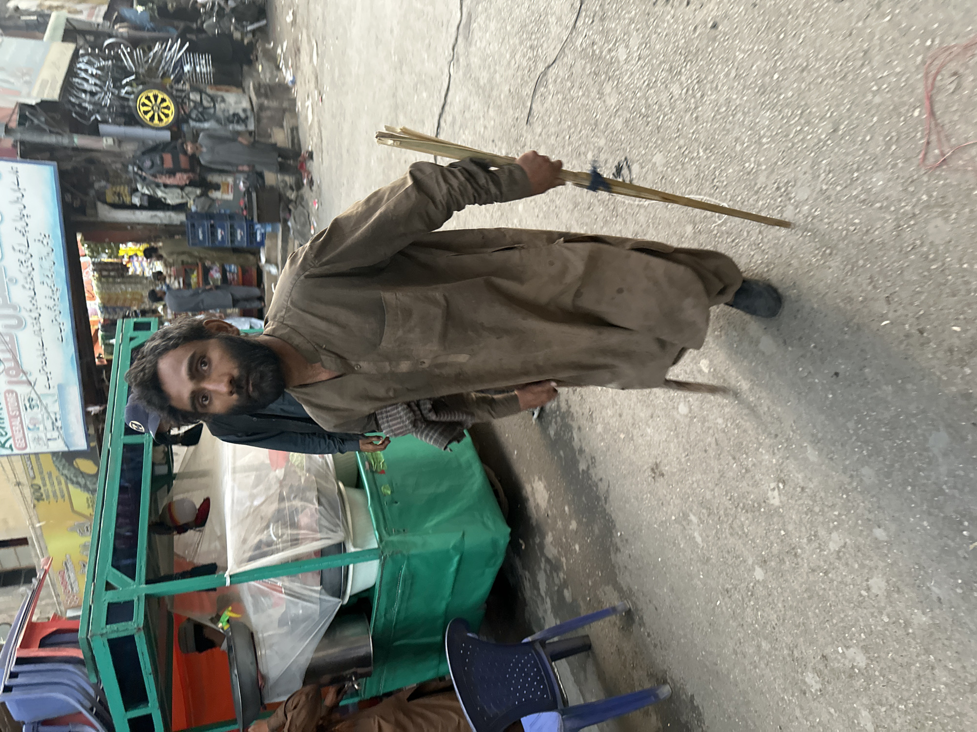 A man stands on a street in Quetta City Tehsil, Pakistan. He has dark hair and a beard and is wearing a dirty brown robe. He holds several sticks tied together in his right hand. To his left is a green metal cart with transparent plastic covering the contents. A blue plastic chair sits near the cart. In the background, there is a shop selling what appears to be motorcycle parts. People are visible in the distance. The street is paved.