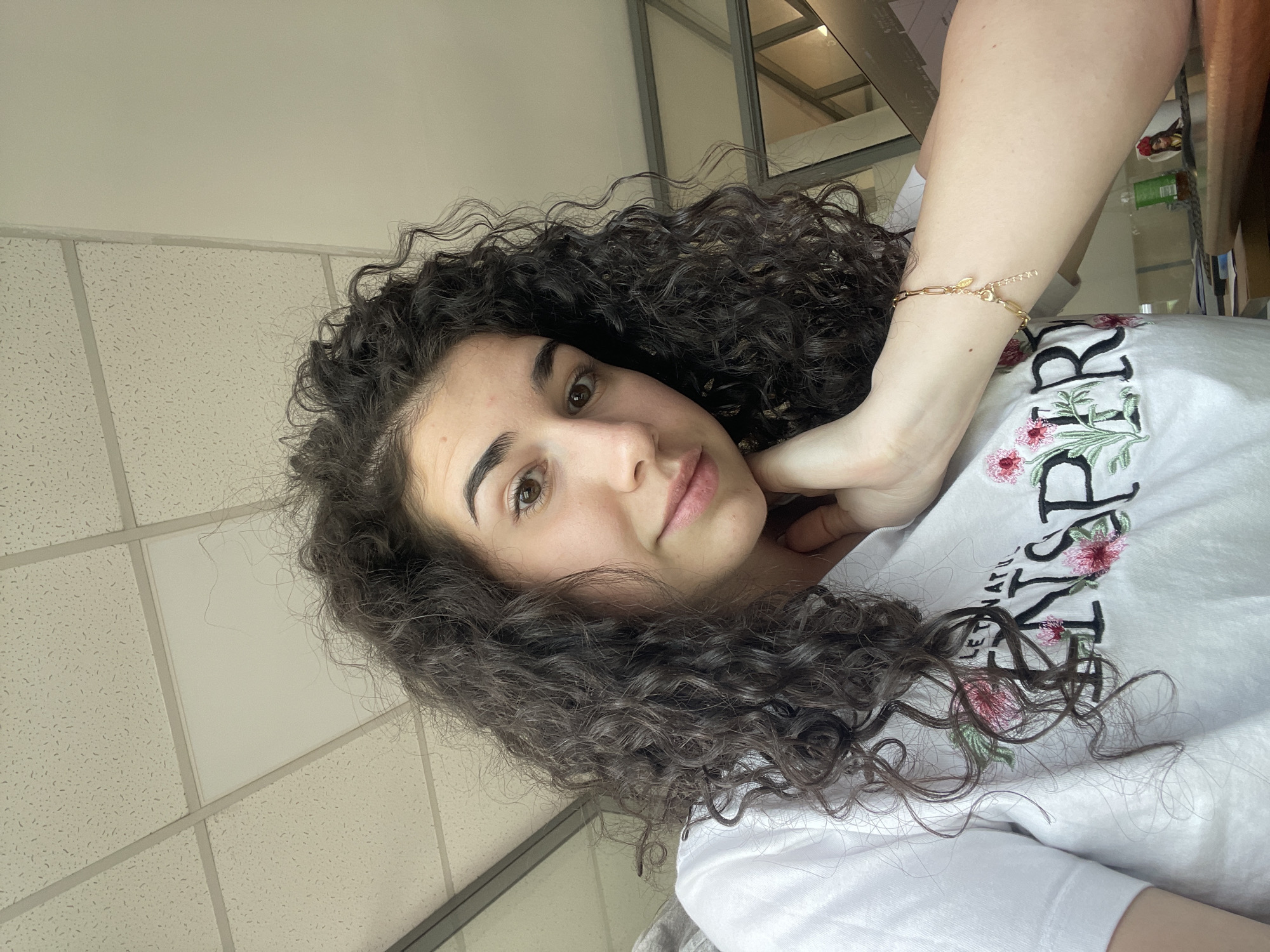 A young woman with dark curly hair is in the photo. She's fair-skinned and is wearing a white t-shirt with floral embroidery. She has a gold bracelet on her right wrist and appears to be resting her chin on her hand. The background shows a ceiling with a grid pattern, commonly found in commercial buildings. It is not possible to ascertain that the location is in Sofia, Bulgaria, based on the information provided in the image.
