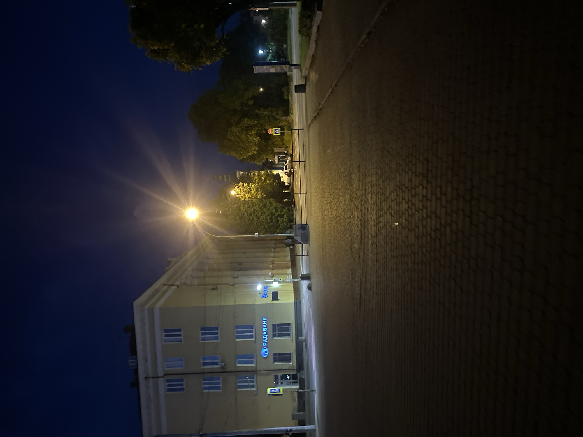 The image depicts a nighttime urban street scene, likely located at or near Voli Square (Площа Волі), 1, Ternopil, Ukraine, identifiable by the "РАДАБАНК" (RADABANK) signage on the primary building and the visible tower of what appears to be the Dominican Church (Кафедральний собор Непорочного Зачаття Пресвятої Діви Марії) in the background.
The sky is dark blue, indicating night. A bright streetlamp in the center-left foreground emits a prominent starburst flare, serving as the main light source. The foreground consists of a dark, textured paved surface, likely cobblestones or interlocking pavers, extending from the bottom of the frame towards the mid-ground.
To the left, a multi-story building, appearing light yellow or beige, features numerous rectangular windows with white or light-colored frames. On its ground floor, a blue, illuminated sign displays "РАДАБАНК" in Cyrillic lettering. Adjacent to the building's entrance, a blue square sign with a white triangle depicting a pedestrian indicates a pedestrian crossing.
In the mid-ground, a road runs horizontally across the frame. Black metal barriers separate the paved foreground from the road. Across the road, a yellow rectangular sign with a red circular 'No Entry' or 'Stop' symbol is visible. Further back, obscured by trees, the top of a distinct dark tower with architectural detailing is discernable. A white sedan is partially visible on the road to the left of this sign.
The right side of the image is dominated by dense, dark green foliage of large trees. Another streetlamp is faintly visible further down the street on the right. A dark cylindrical trash receptacle stands near the curb, and a tall, dark rectangular signpost or kiosk is positioned further along the sidewalk on the right. No individuals are visibly present in the scene.