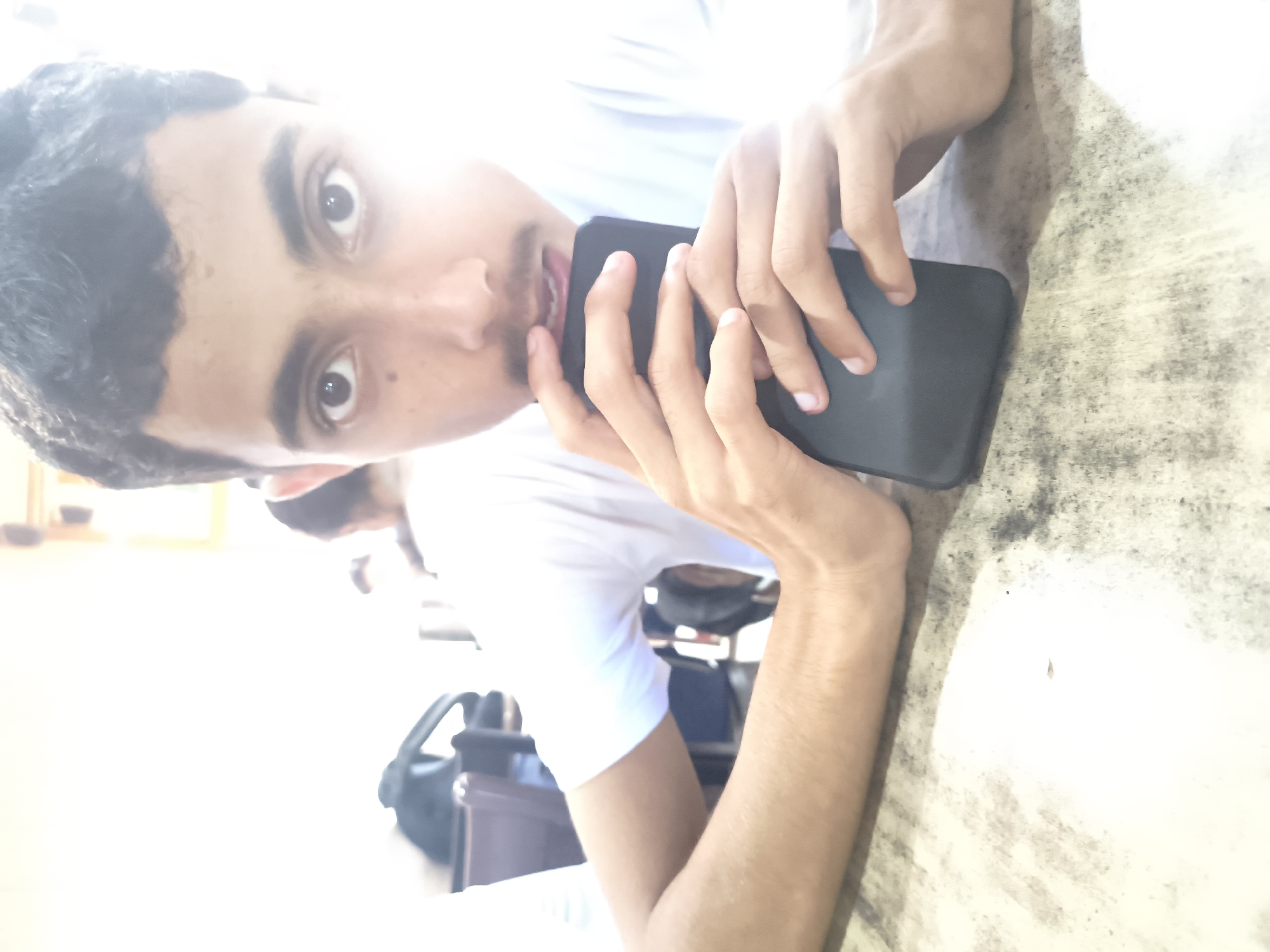 A young man, appearing to be in his late teens or early twenties, is shown close-up, holding a dark-colored mobile phone in front of his face. He is wearing a white t-shirt. His hands are positioned as if he is about to use or is currently using the phone. The background is blurred and appears to be an indoor setting with a table in the foreground. The lighting is bright, causing some overexposure, particularly around the man's head and the right side of the image. The location is identified as Dharamshala, India.