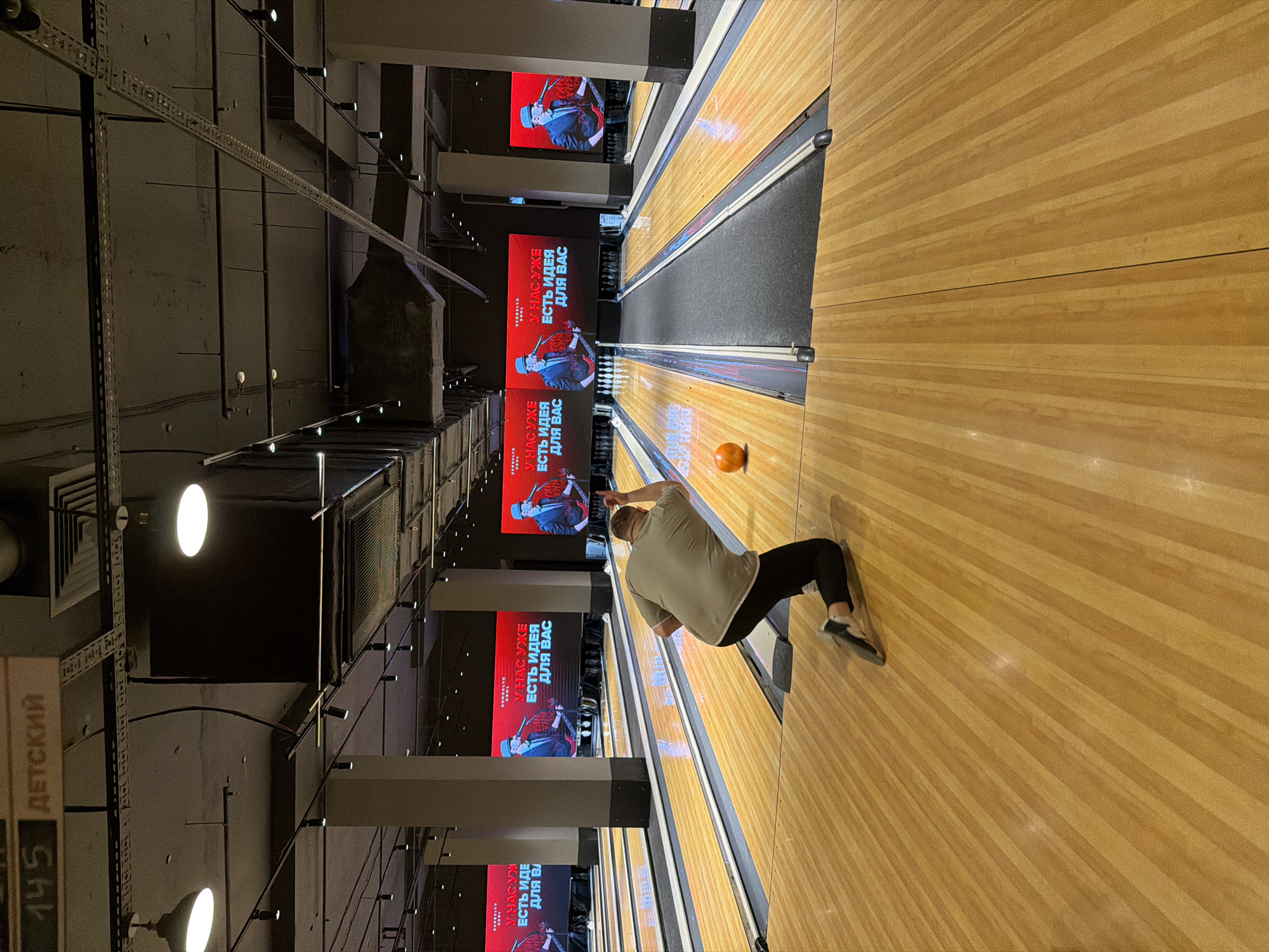 The image shows a man bowling in what appears to be a bowling alley. He is in the middle of his swing, releasing an orange bowling ball. He is wearing a light-colored t-shirt, black pants, and bowling shoes. The bowling lane is made of wood with dark grey gutters. Several large screens hang above the lanes displaying promotional content in Russian. The text on the screens reads "У НАС УЖЕ ЕСТЬ ИДЕЯ ДЛЯ ВАС", which translates to "WE ALREADY HAVE AN IDEA FOR YOU". The location, Izhevsk, Russia, is indicated by the text on one of the signs, which includes the word "ДЕТСКИЙ", commonly seen on signs in Russia. The alley is indoors, with industrial-style lighting and ceiling fixtures.