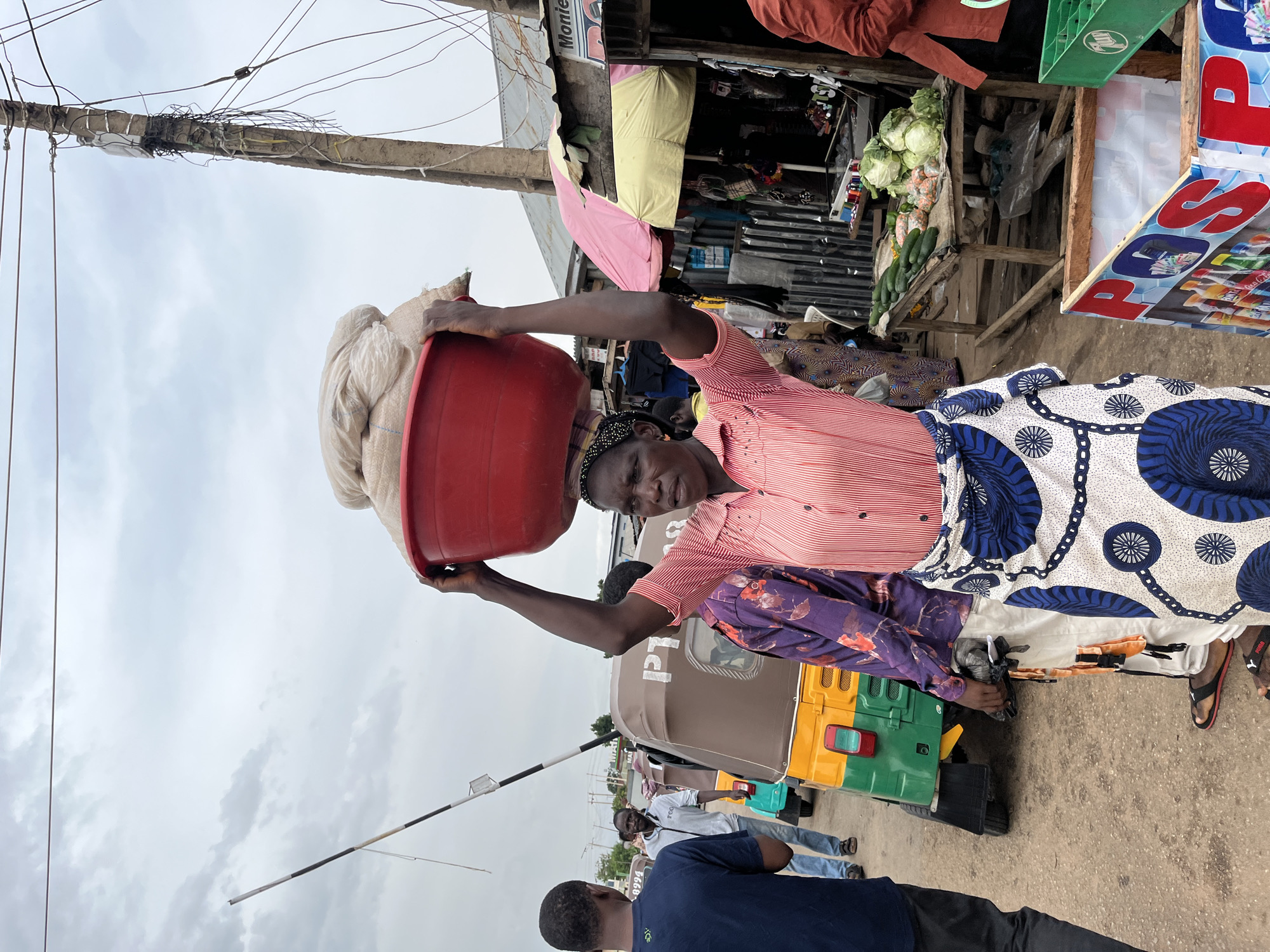 The original image, if viewed upright, captures a dynamic street scene in Gura Top, Nigeria, likely a market or commercial area, under an overcast sky. The central figure is a woman with dark skin, positioned slightly off-center, balancing a large, full red plastic basin on her head. She uses her left hand to steady the basin. She wears a light red and white striped short-sleeved button-down shirt, a dark blue and white patterned ankle-length skirt, and a dark head covering with a light band. A purple patterned garment is visible between her arm and torso.
To the right and behind the woman, several makeshift market stalls are visible. One stall displays fresh produce, including neat piles of green cabbages and cucumbers. Another stall features a yellow and pink awning and a prominent blue and red sign with "POS" written in large letters. Above these stalls, a partial sign reads "Monte...".
Several other people are present in the scene. A brown and green Keke Napep (auto-rickshaw) is visible behind the main woman, with "IPT" written on its side. In the foreground, facing away from the camera, a man in a dark blue t-shirt with "ACE" printed on the back is visible. Other individuals are scattered in the background, interacting or moving within the bustling environment.
The sky is cloudy and grey, suggesting daytime. A utility pole, seemingly old and burdened with numerous wires, stretches across the upper left portion of the frame. A black and white striped barrier arm extends diagonally from the mid-ground towards the lower left, possibly indicating an entry point or checkpoint. The ground is a mix of dirt and rough pavement. The number "8994" is faintly visible on the lower left side of the image. The scene portrays daily life and commerce in a busy street setting.